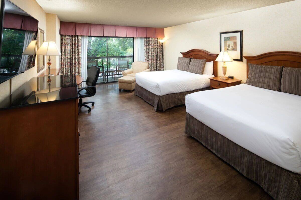 A hotel room with two beds, a desk, a chair, and a balcony with a view of trees.