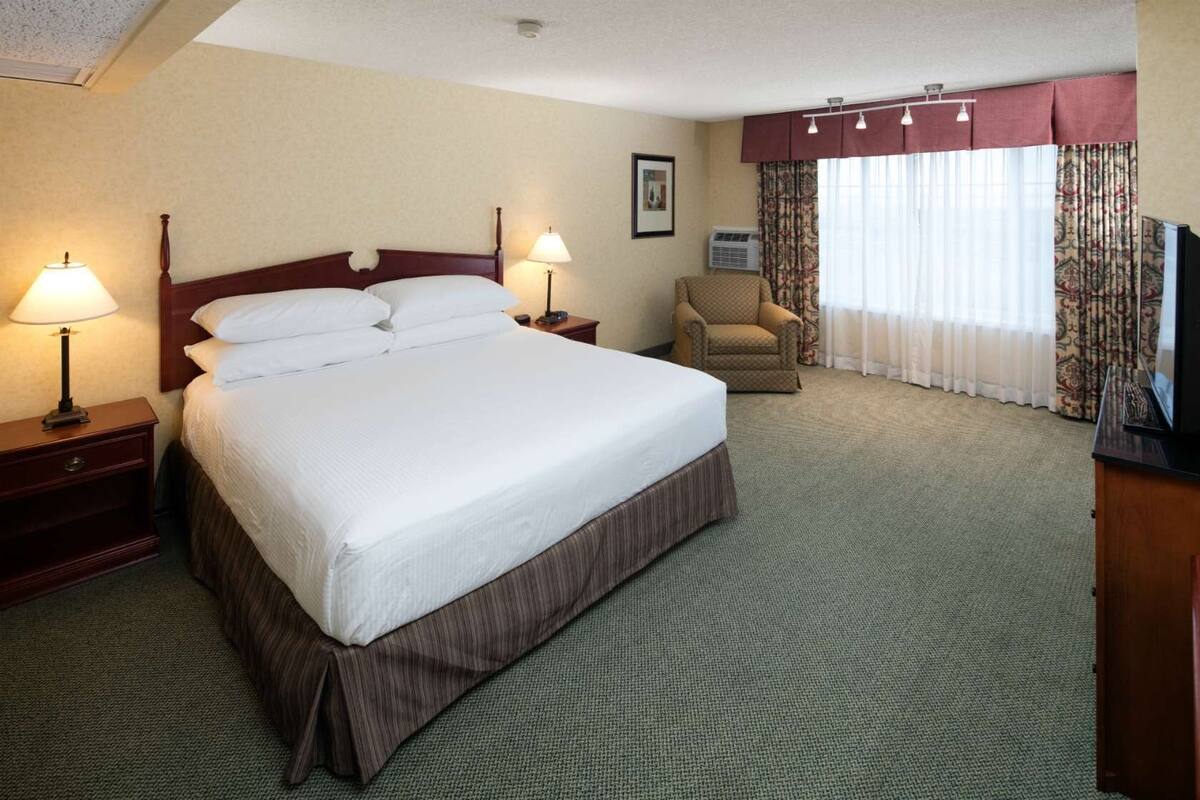 A hotel room with a large bed, two bedside tables with lamps, a chair, a TV, and a window with curtains.