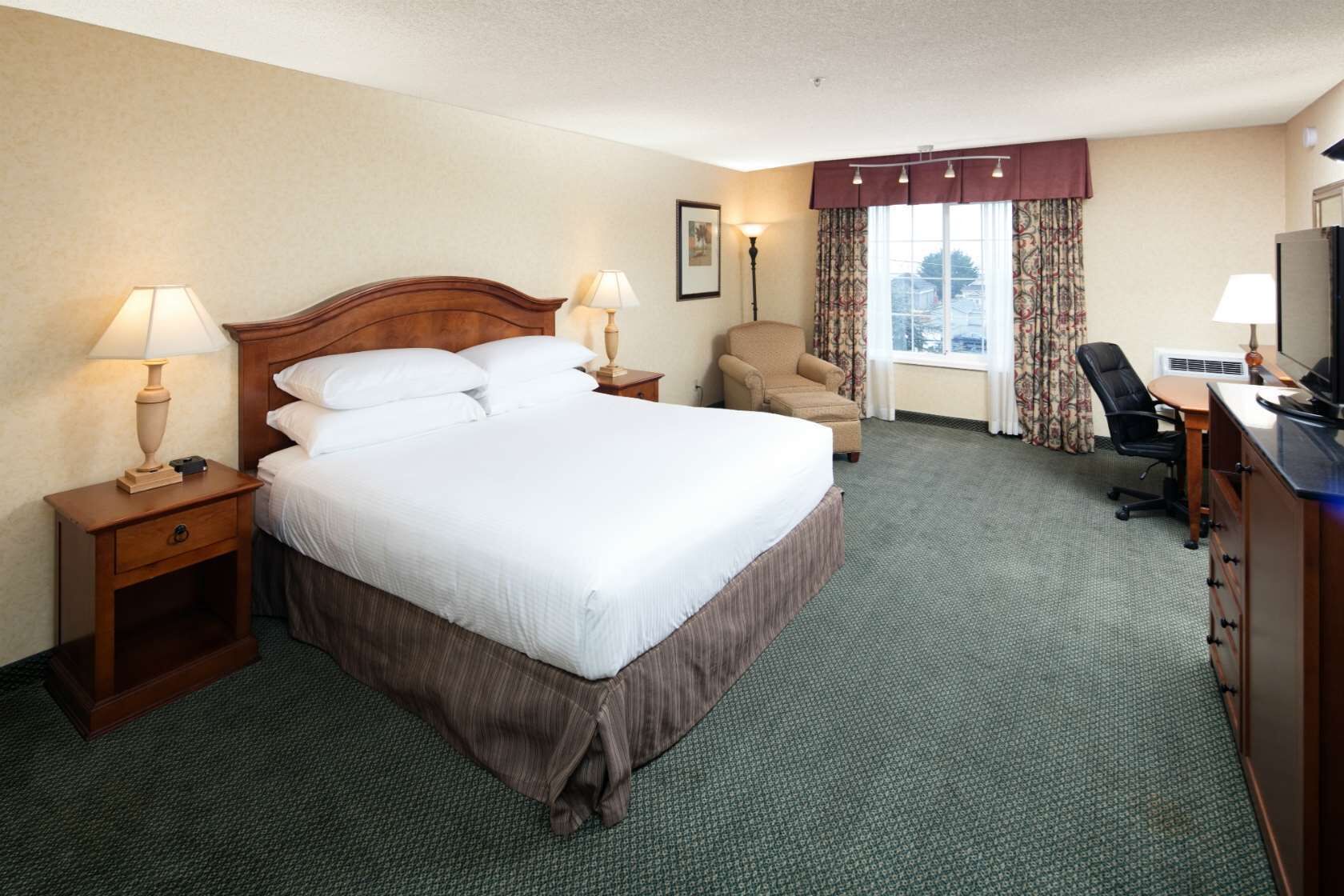 A hotel room with a large bed, two bedside tables with lamps, a chair, a desk with a chair, and a window with curtains.