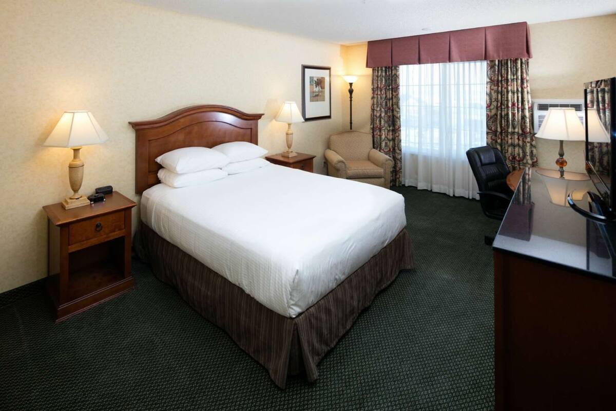 A hotel room with a large bed, two bedside tables with lamps, a chair, a desk with a mirror, and a window with curtains.
