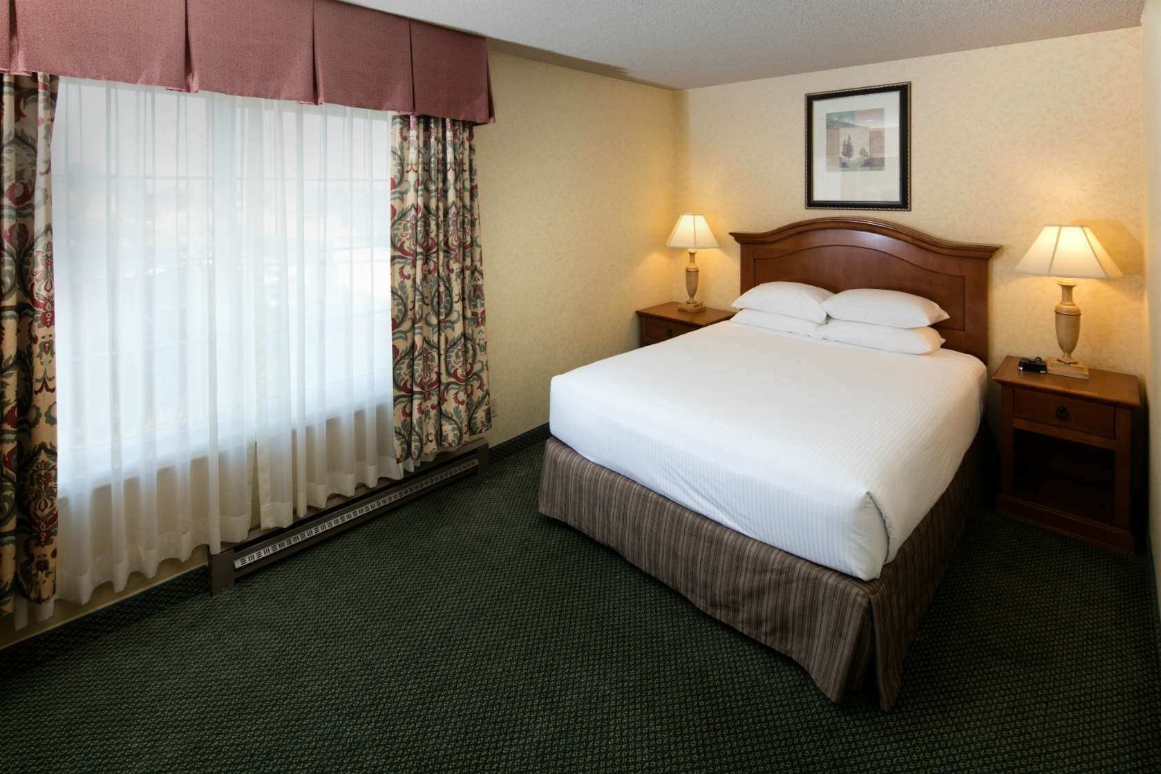 Una habitación de hotel con una cama grande, dos mesitas de noche con lámparas, una ventana con cortinas traslúcidas y cortinas decoradas, y un cuadro en la pared.