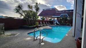 Una piscina con una escalera metálica, sillas de descanso, palmeras y un edificio con techo rojo al fondo.