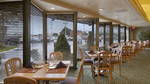 Un restaurante con varias mesas y sillas, amplias ventanas con persianas y vista al estacionamiento.