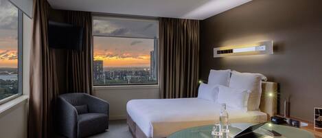 A hotel room with a bed, a glass table with a laptop, a chair, a TV, and a view of the city at sunset.