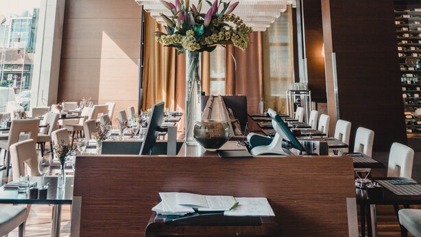 Un interior de restaurante moderno con paneles de techo de madera, un arreglo floral central y vistas de la ciudad a través de amplias ventanas.