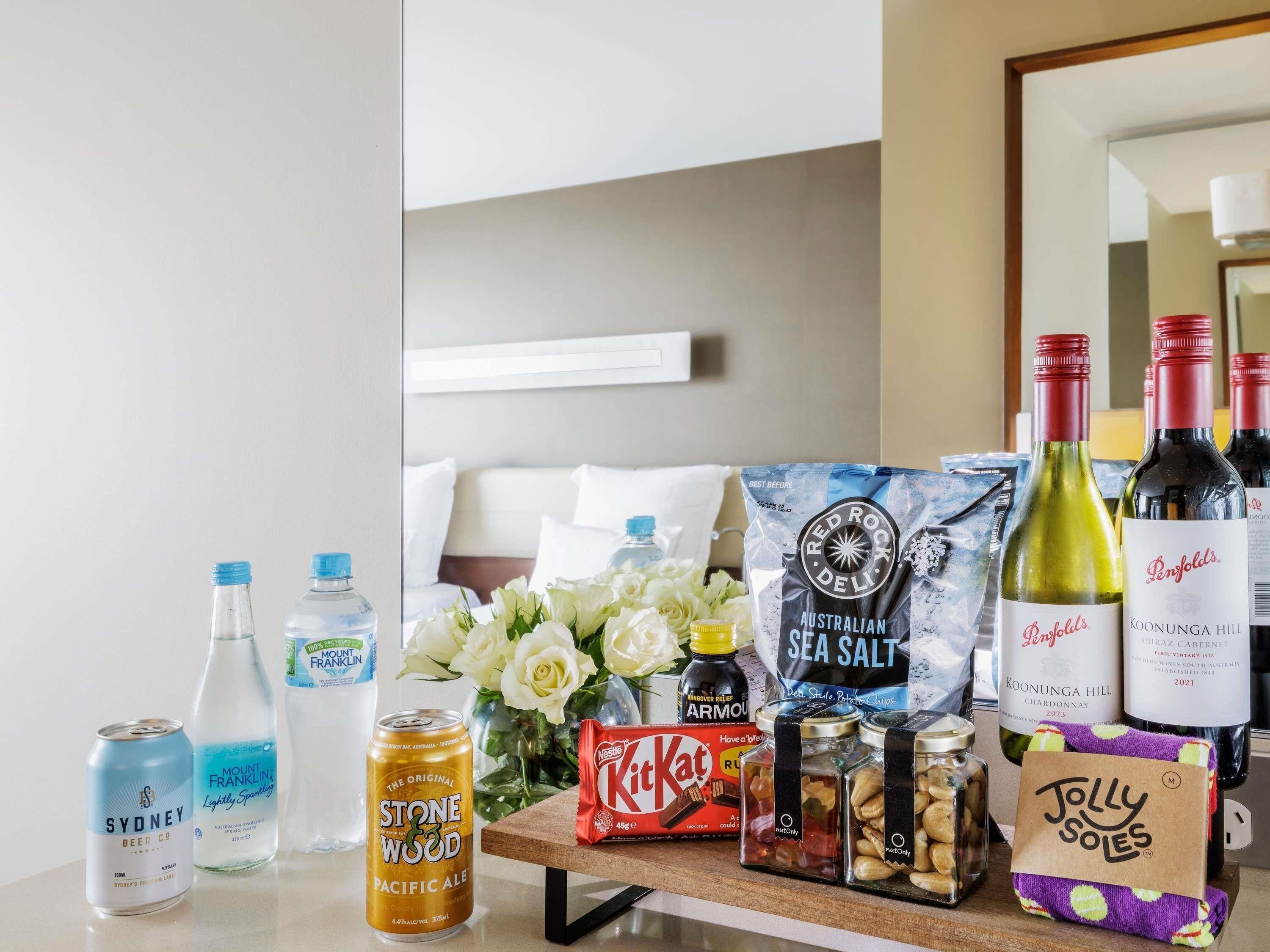 Una habitación de hotel con una cama, una mesita de noche con diversos objetos, incluyendo botellas de vino, aperitivos y una botella de agua.