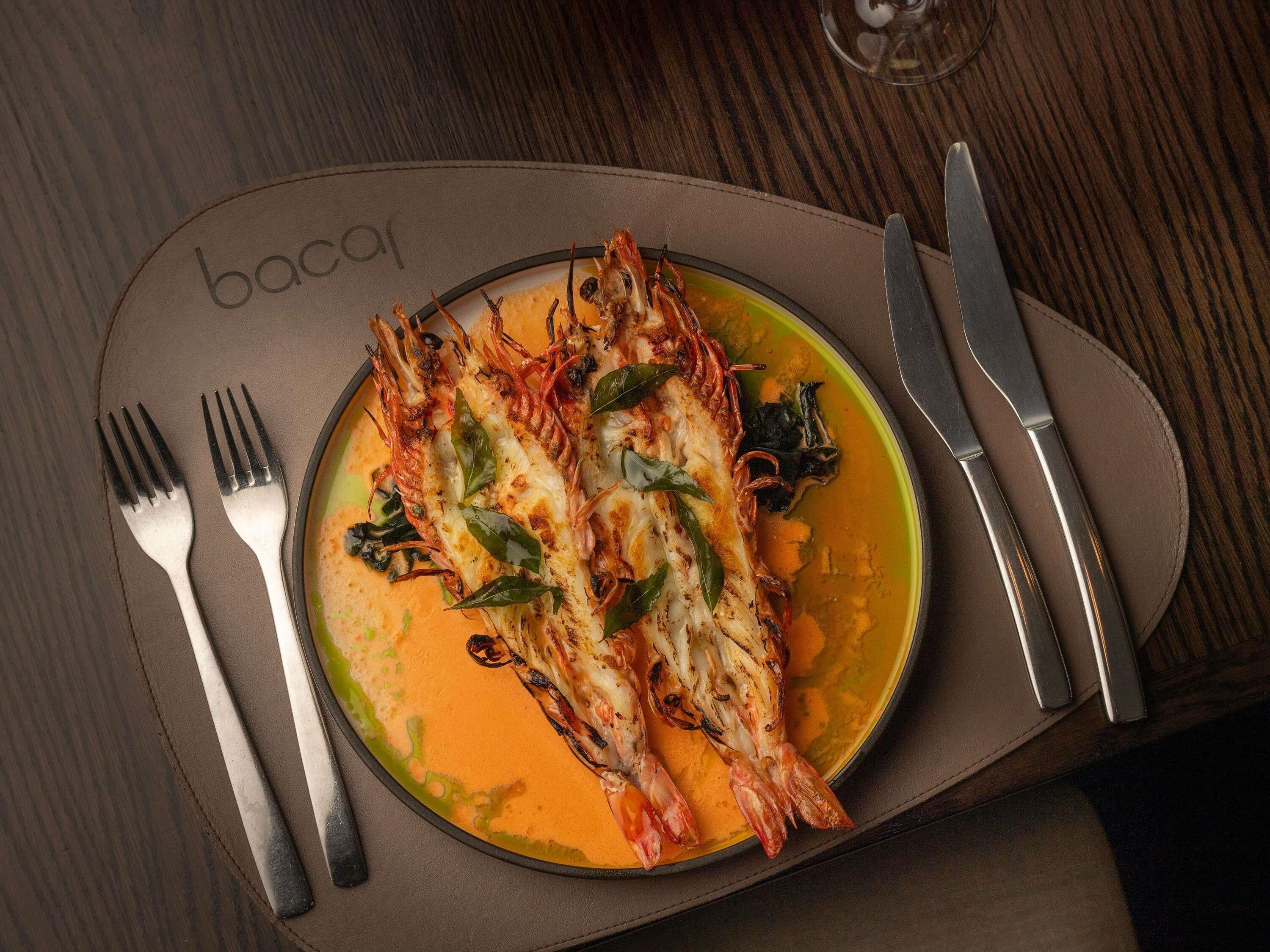 A plate of grilled shrimp with herbs, accompanied by a fork and knife on a brown placemat with the word 'bacai' on it.