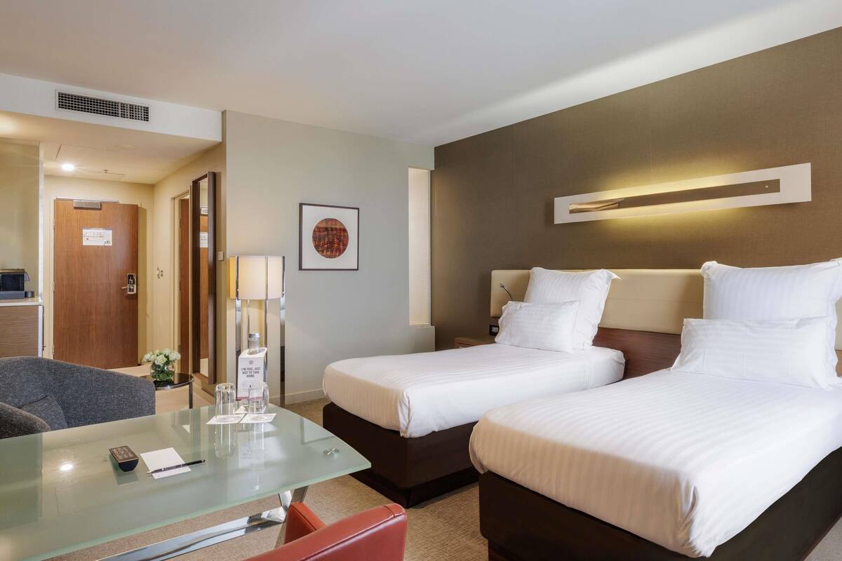 Habitación de hotel con dos camas, una mesita de vidrio con un control remoto, una lámpara y un cuadro en la pared.
