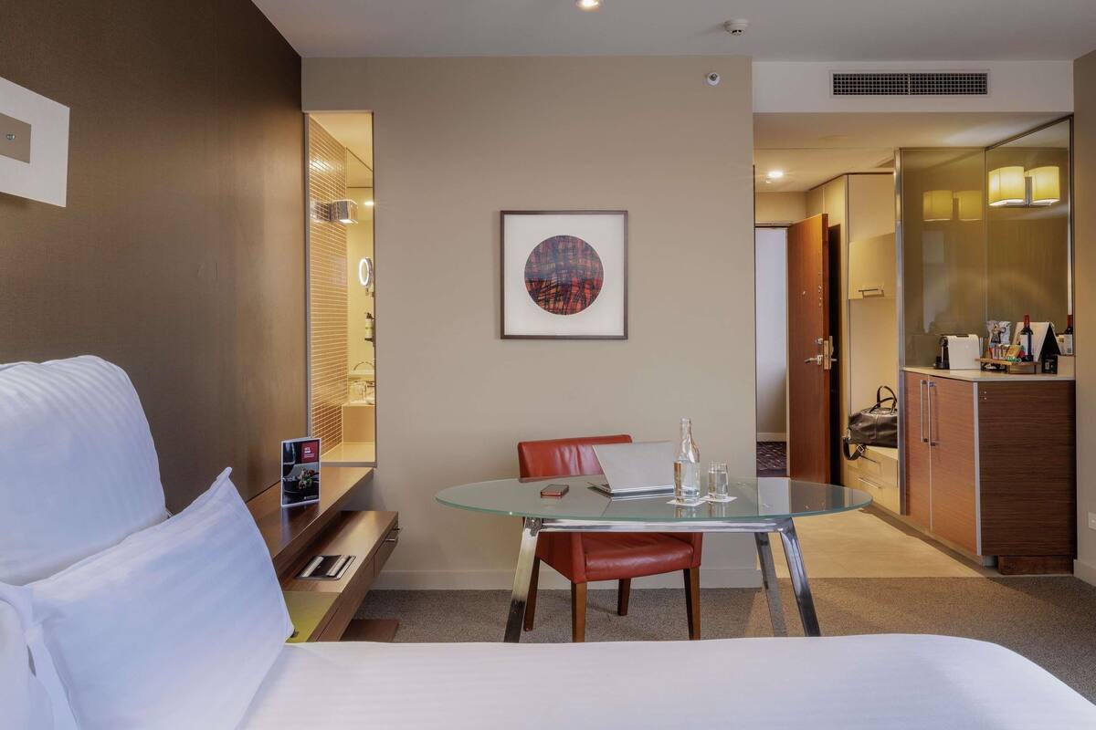 Habitación de hotel con una cama, una mesa de vidrio con sillas, un escritorio pequeño con una computadora portátil, un espejo y un cuadro en la pared.