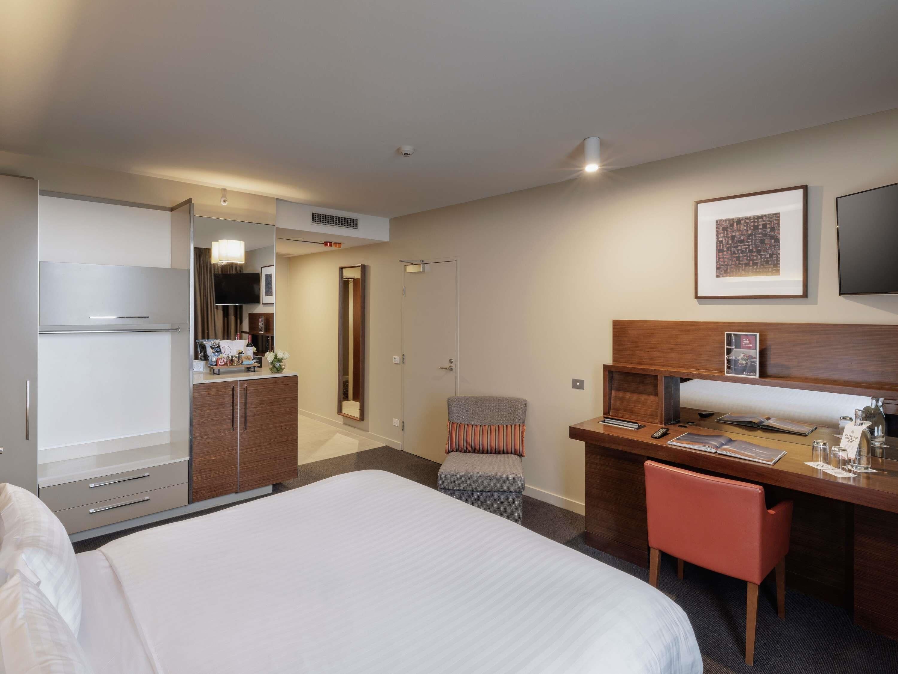 Quarto de hotel com cama, escrivaninha, cadeira, televisão, espelho e porta para o banheiro.