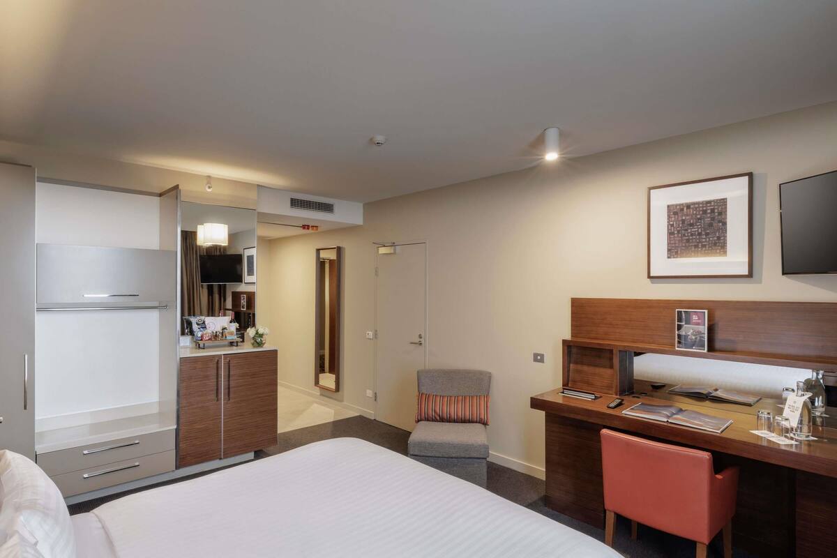 Un cuarto de hotel con cama, escritorio con silla, televisión, espejo y puerta que da a un baño.