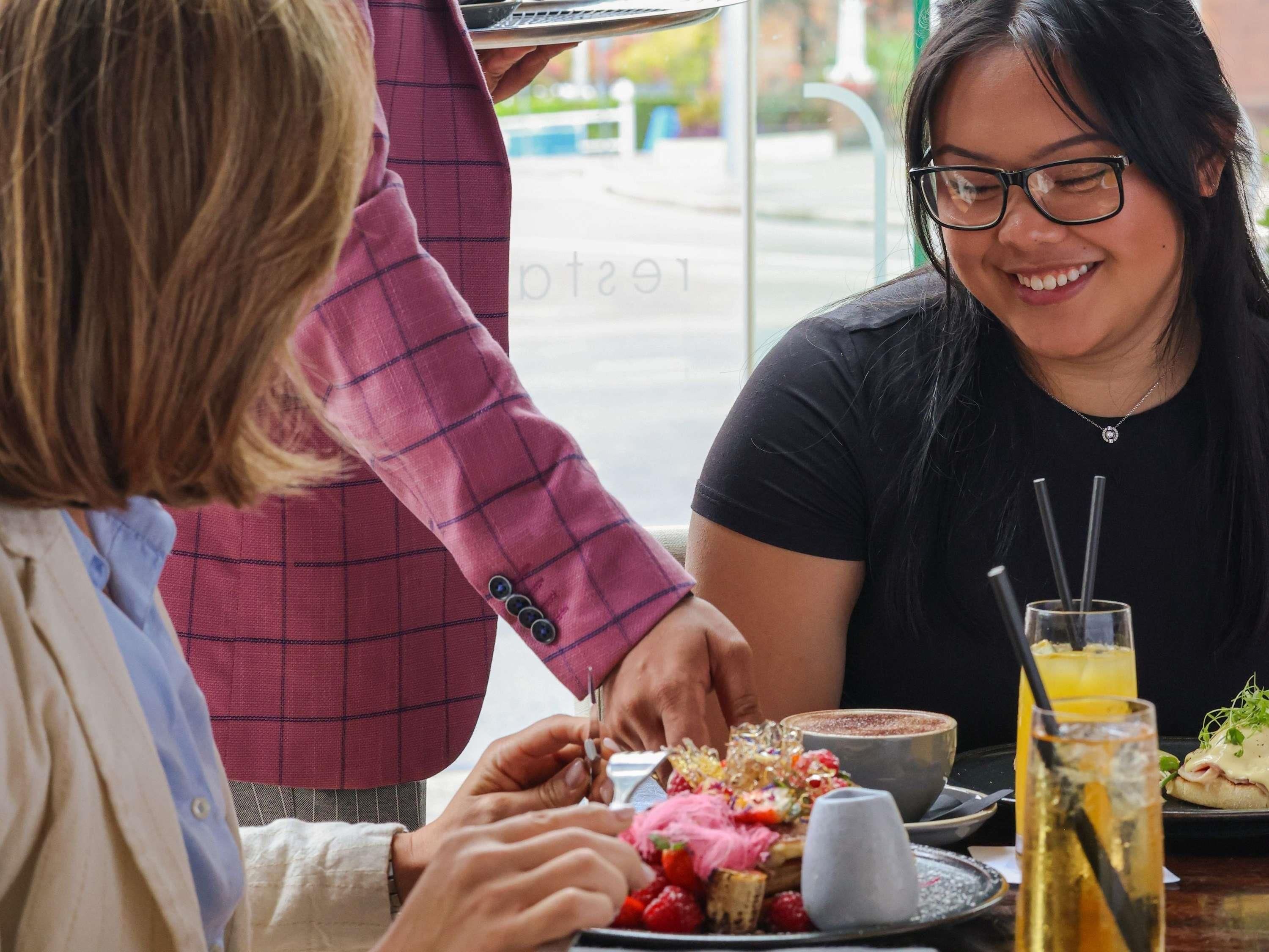 A person serving food at a table with a variety of dishes, including a dessert and a beverage.