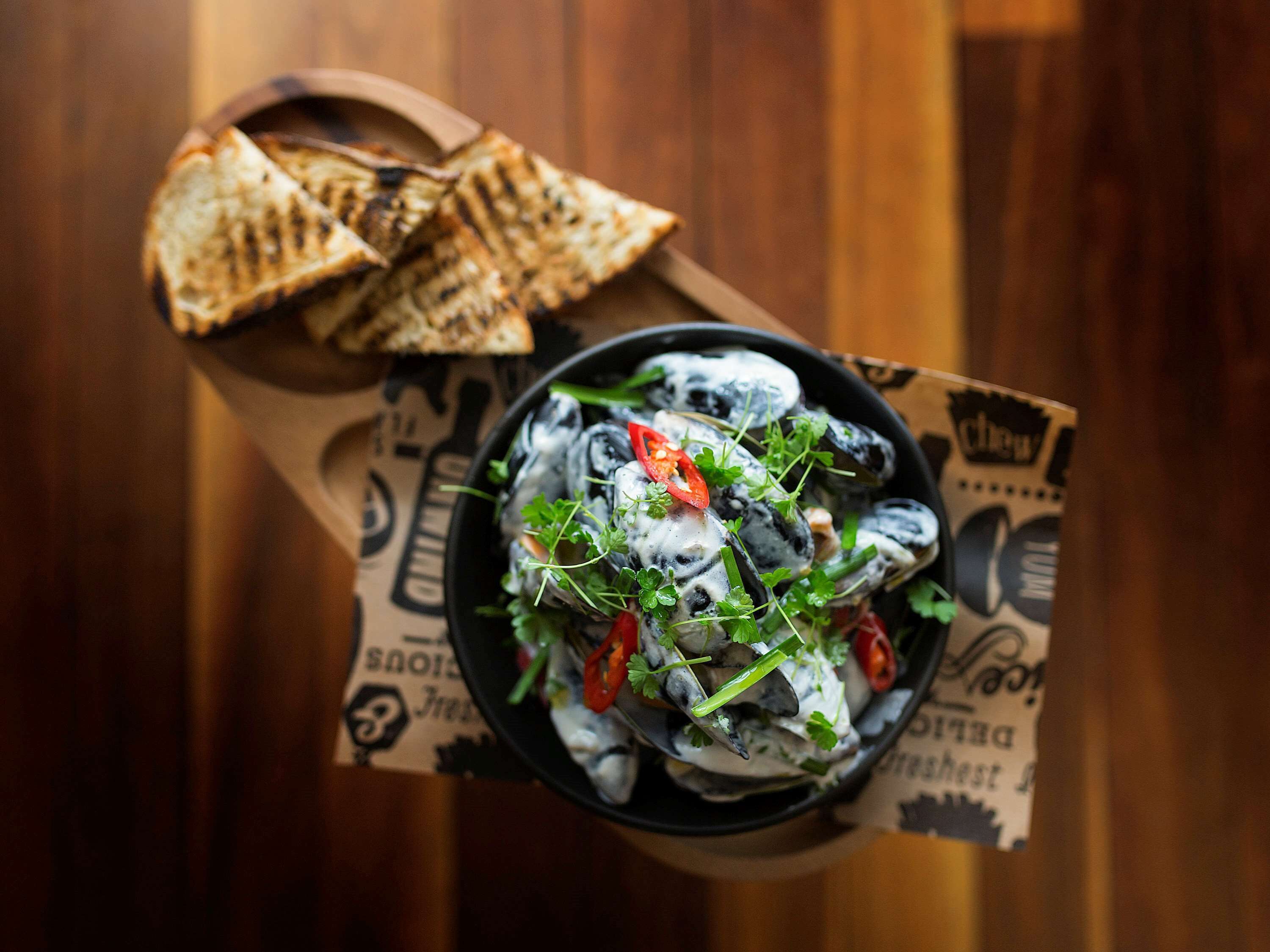 A bowl of food with mussels, garnished with herbs and red chili, on a wooden table with a menu.