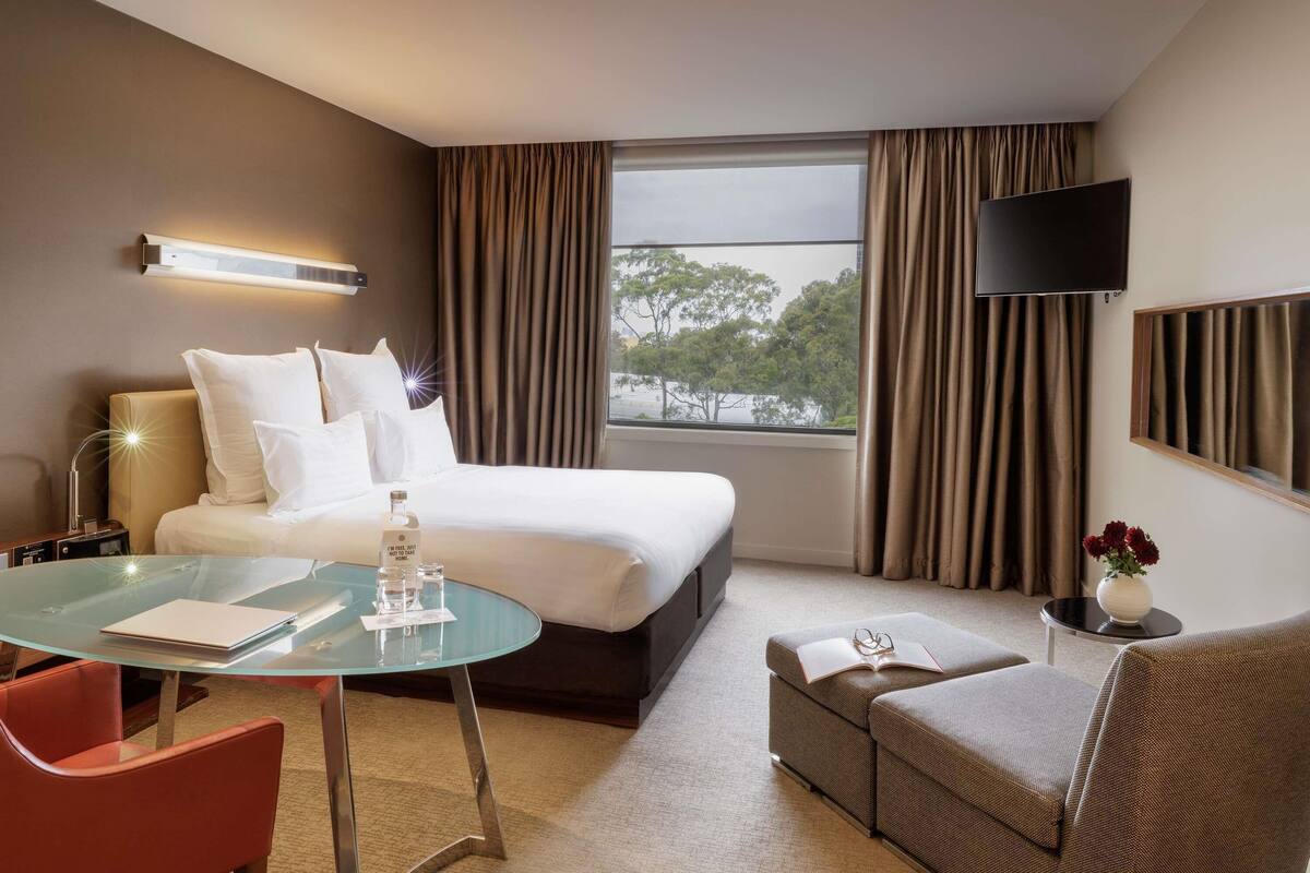 Habitación de hotel con cama, una mesita de cristal, una silla roja, sofá, televisor y ventana con cortinas.