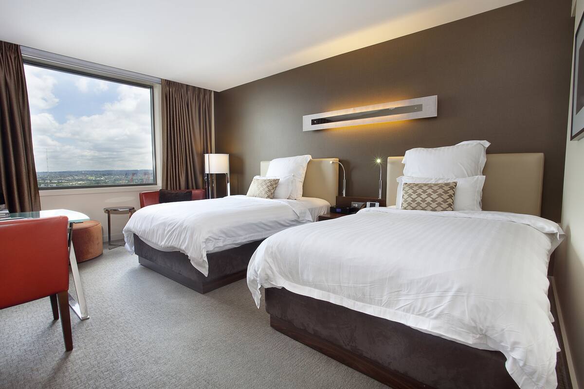 Habitación de hotel con dos camas, un escritorio, una silla y un ventanal con vistas a la ciudad.