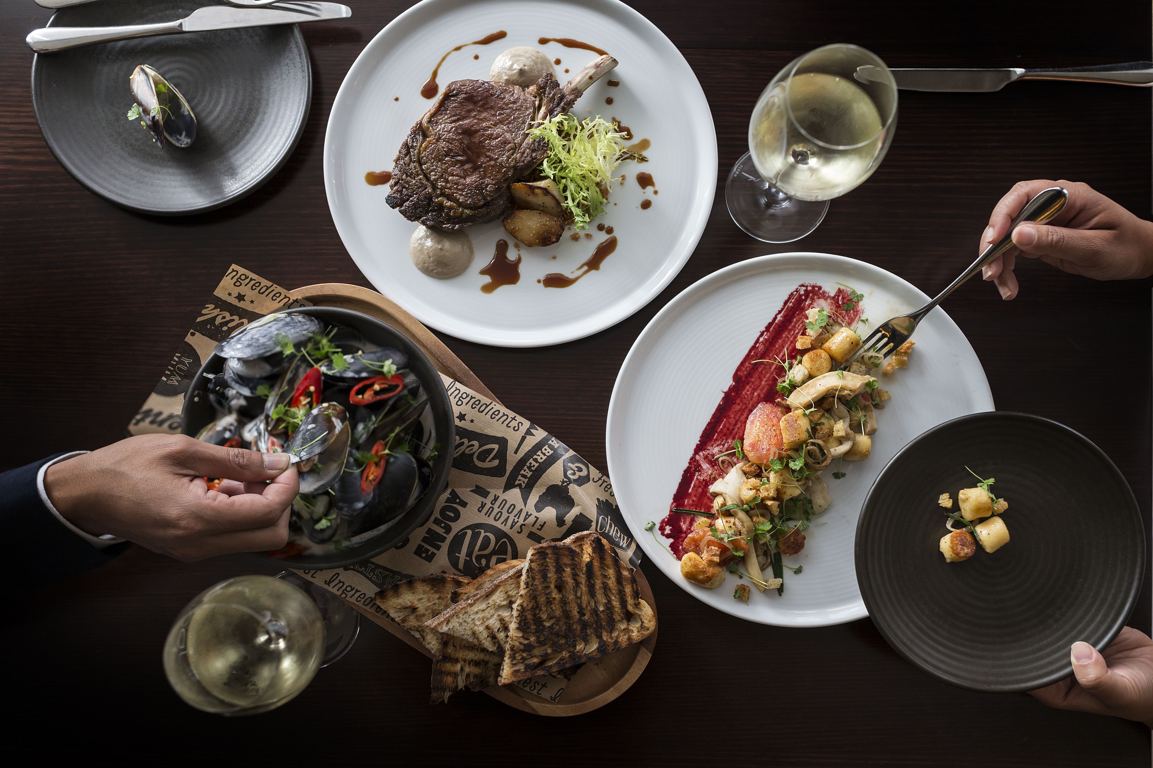 A dining table with various dishes, including steak, vegetables, and bread, along with wine glasses and cutlery.