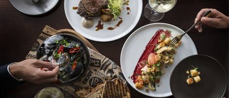 A dining table with various dishes, including steak, vegetables, and bread, along with wine glasses and cutlery.