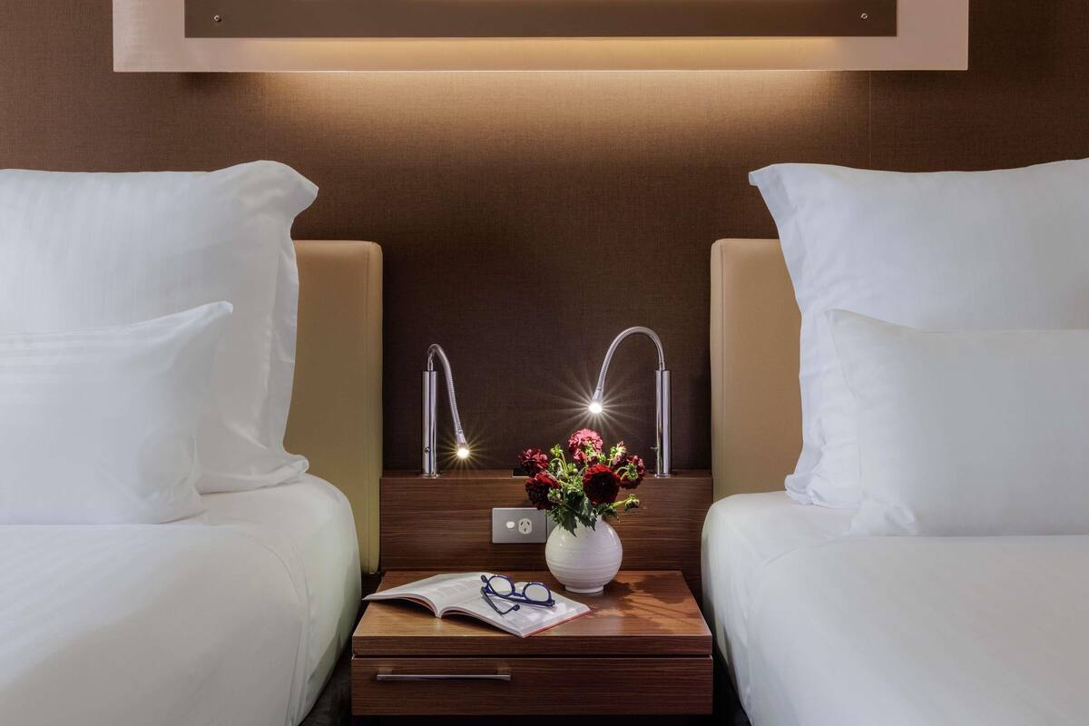 Habitación de hotel con dos camas, una mesita de noche con un florero y una lámpara fijada a la pared.