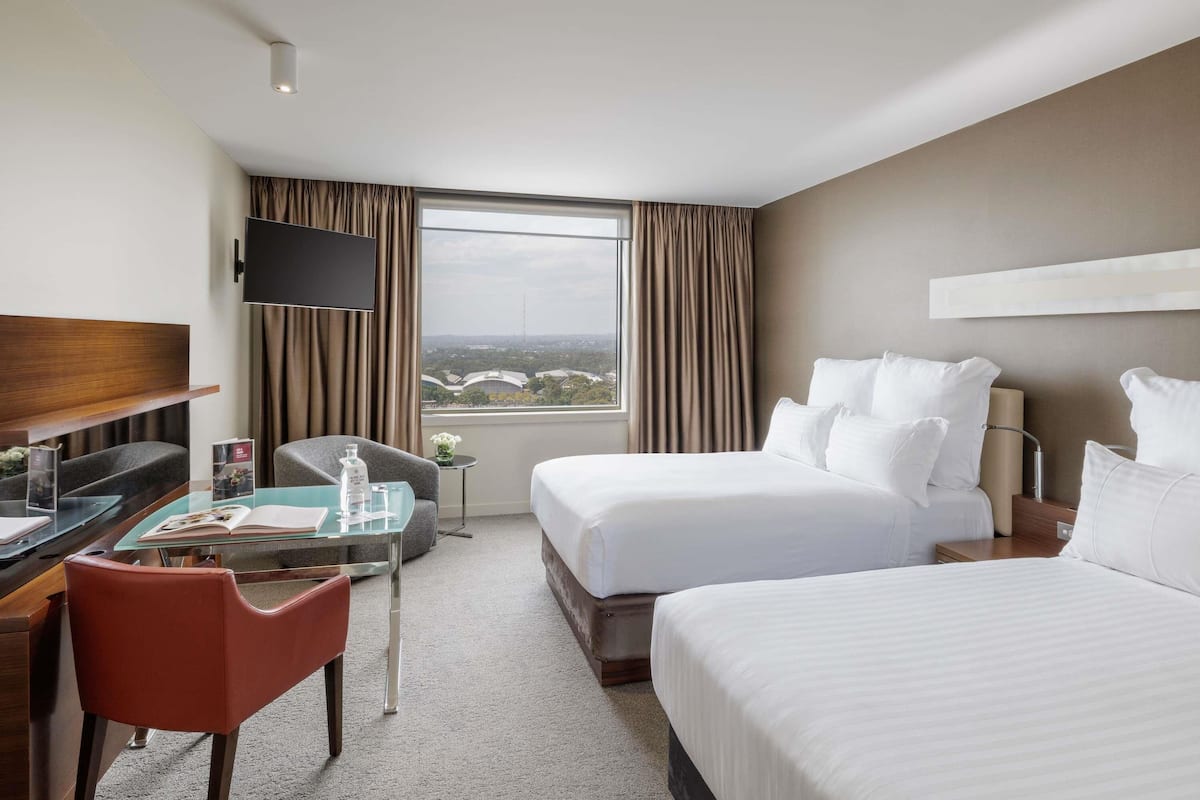A hotel room with a desk, two beds, a TV, and a window with a city view.