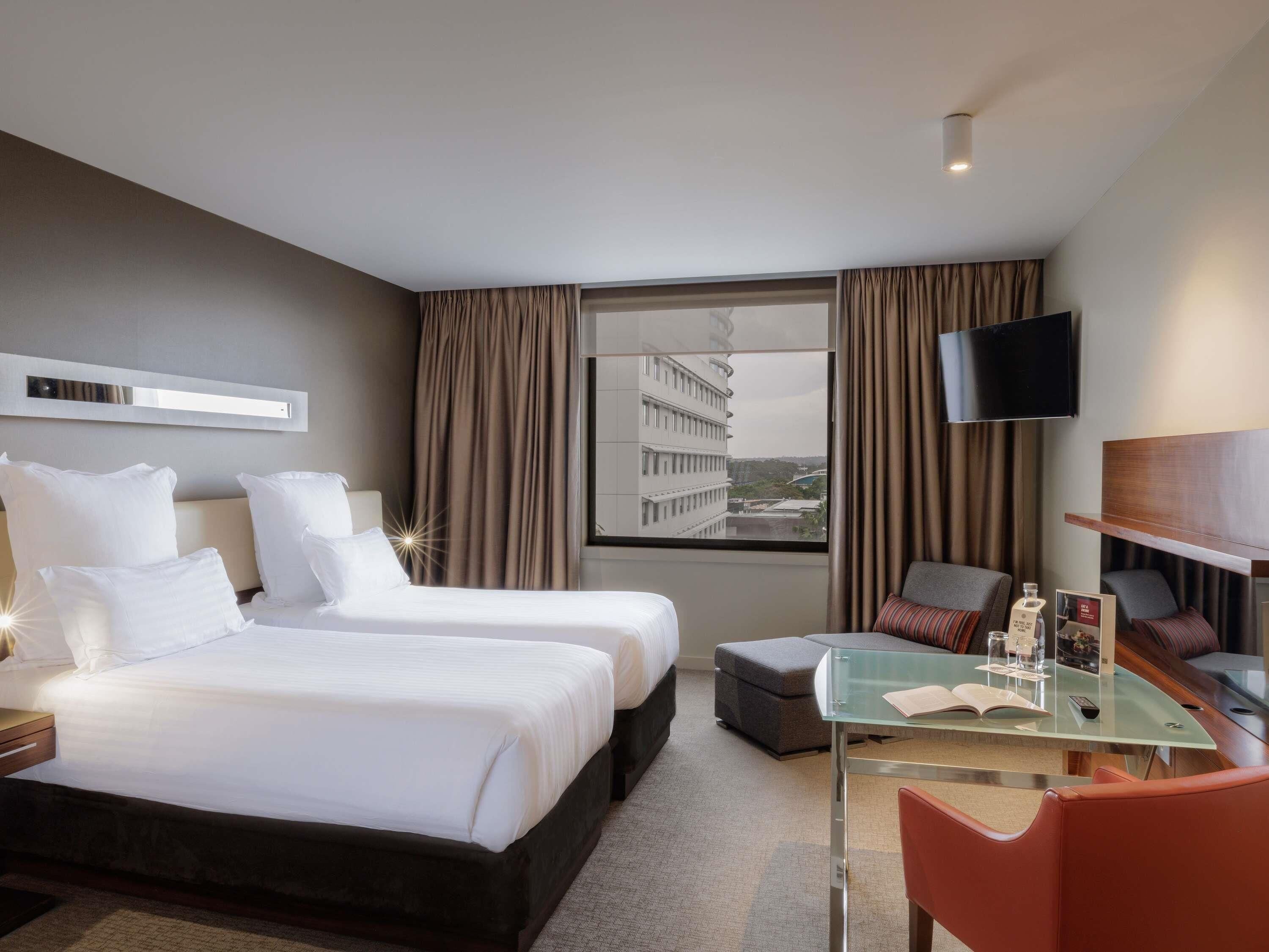Een hotelkamer met twee bedden, een bureau met stoel, een televisie en een groot raam met uitzicht op gebouwen.