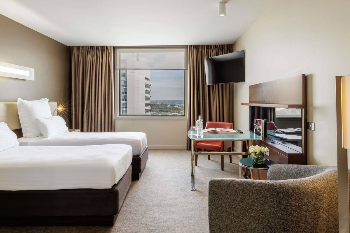 Habitación de hotel con dos camas, un escritorio, un televisor y vista a un paisaje urbano.