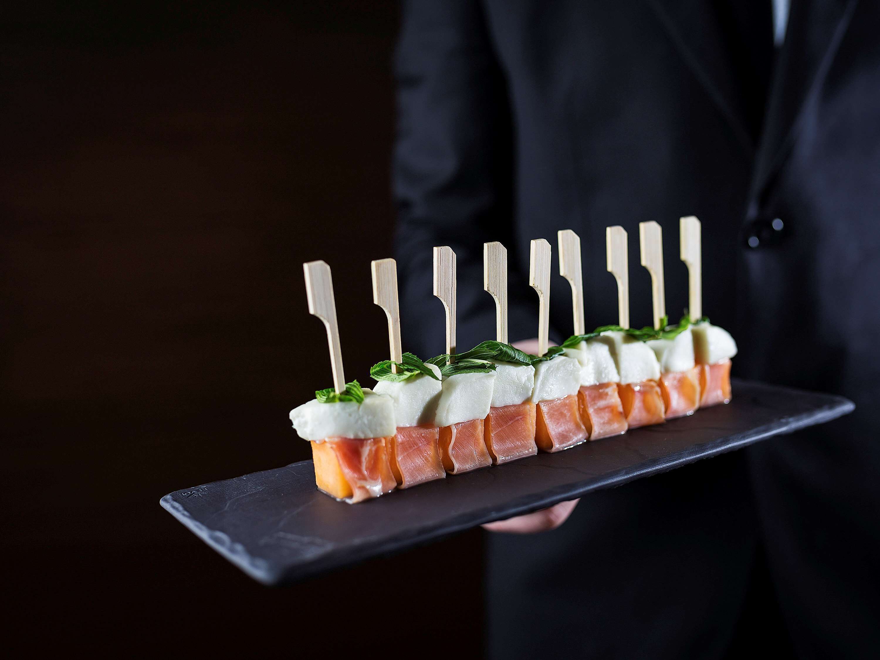 A person holding a tray with a food arrangement consisting of alternating slices of smoked salmon and cream cheese, garnished with mint leaves, and skewered with wooden sticks.