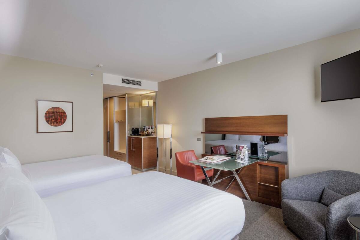 Habitación de hotel con dos camas, un escritorio con silla, un televisor y un cuadro en la pared.