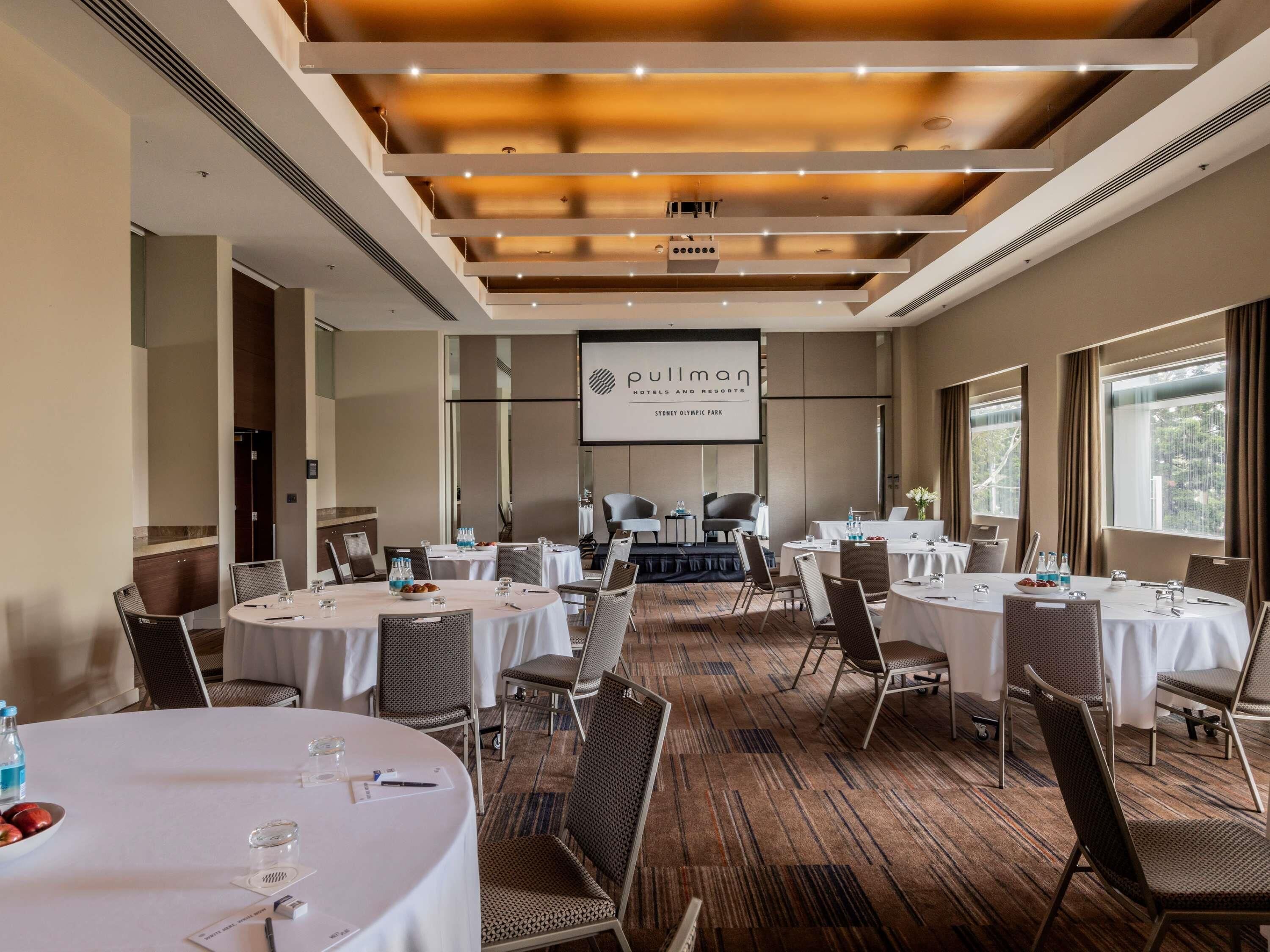 A conference room with round tables, chairs, and a projection screen displaying 'Pullman Hotels and Resorts Sydney Olympic Park'.