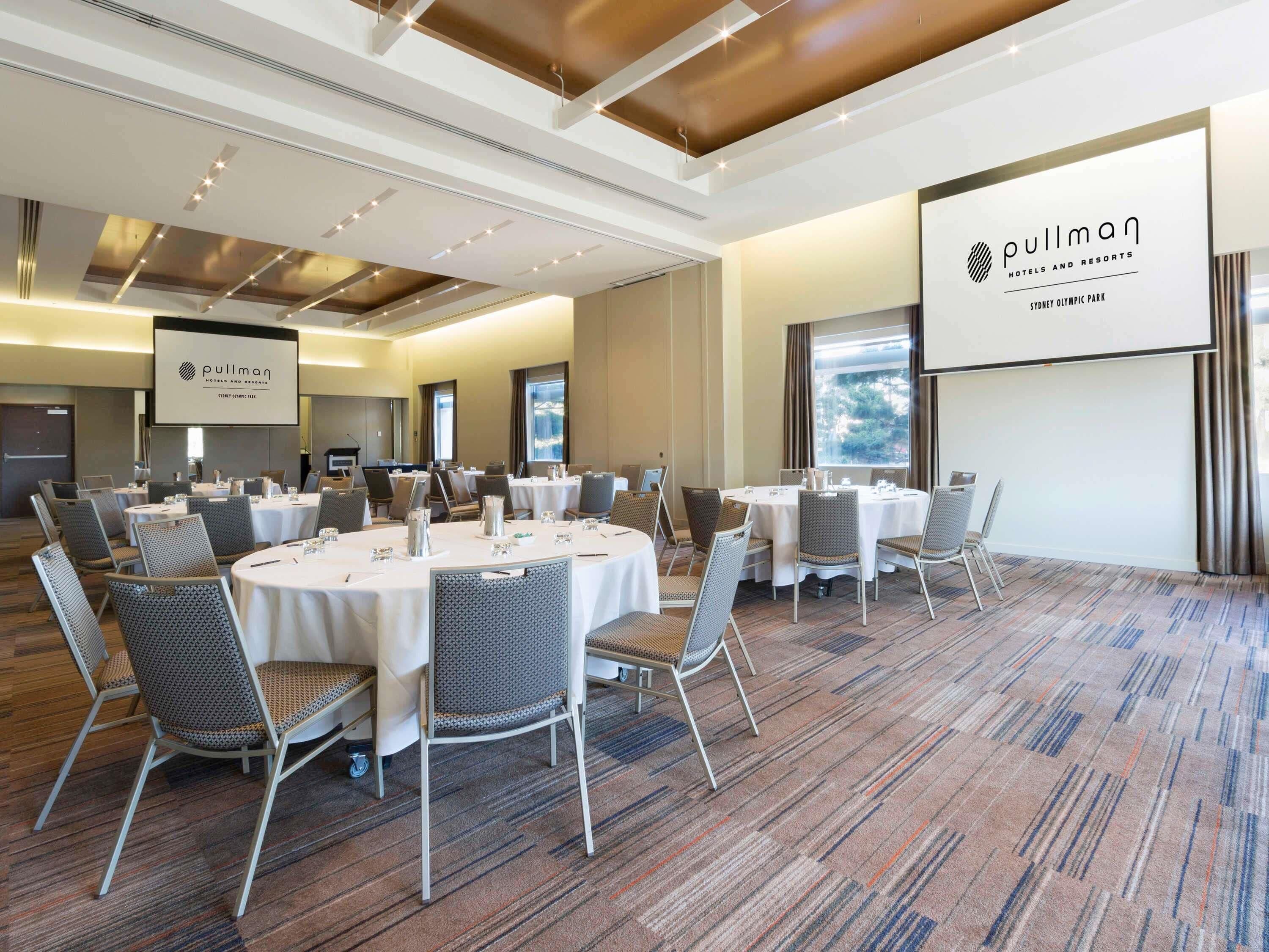 Un salón de conferencias con mesas redondas, sillas y una pantalla con el logo de Pullman.