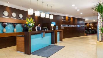 A hotel reception area with a wooden reception desk, multiple clocks, and a digital screen.