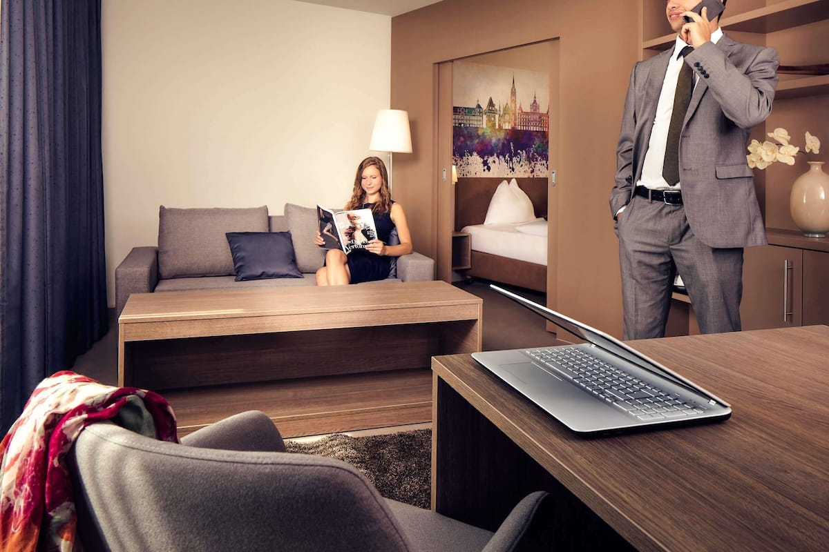 A modern hotel room with a sofa, a bed, a desk with a laptop, and a person talking on the phone.