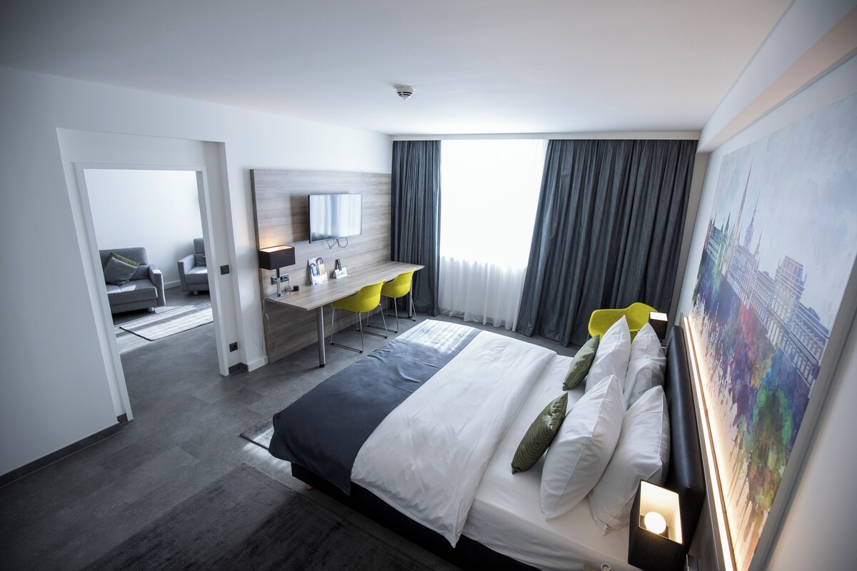 A modern hotel room with a large bed, a desk with chairs, a flat-screen TV, and a cityscape painting on the wall.
