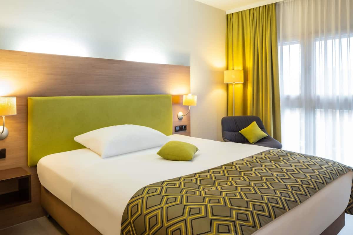 A hotel room with a large bed, a green headboard, a patterned throw blanket, a green pillow, a chair with a yellow pillow, and a window with yellow curtains.