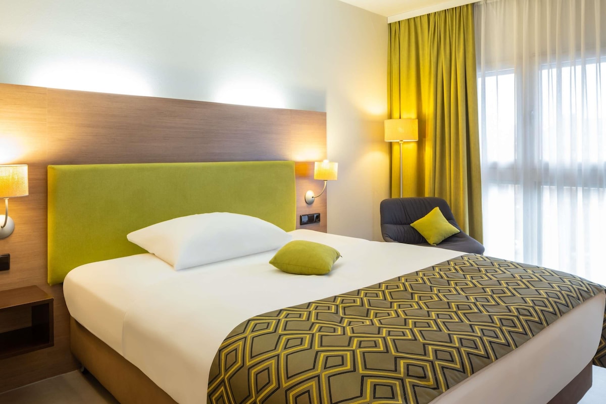 A hotel room with a large bed, a green headboard, a patterned throw blanket, a green pillow, a chair with a yellow pillow, and a window with yellow curtains.