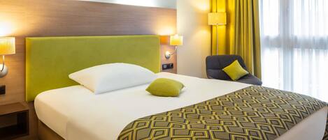 A hotel room with a large bed, a green headboard, a patterned throw blanket, a green pillow, a chair with a yellow pillow, and a window with yellow curtains.