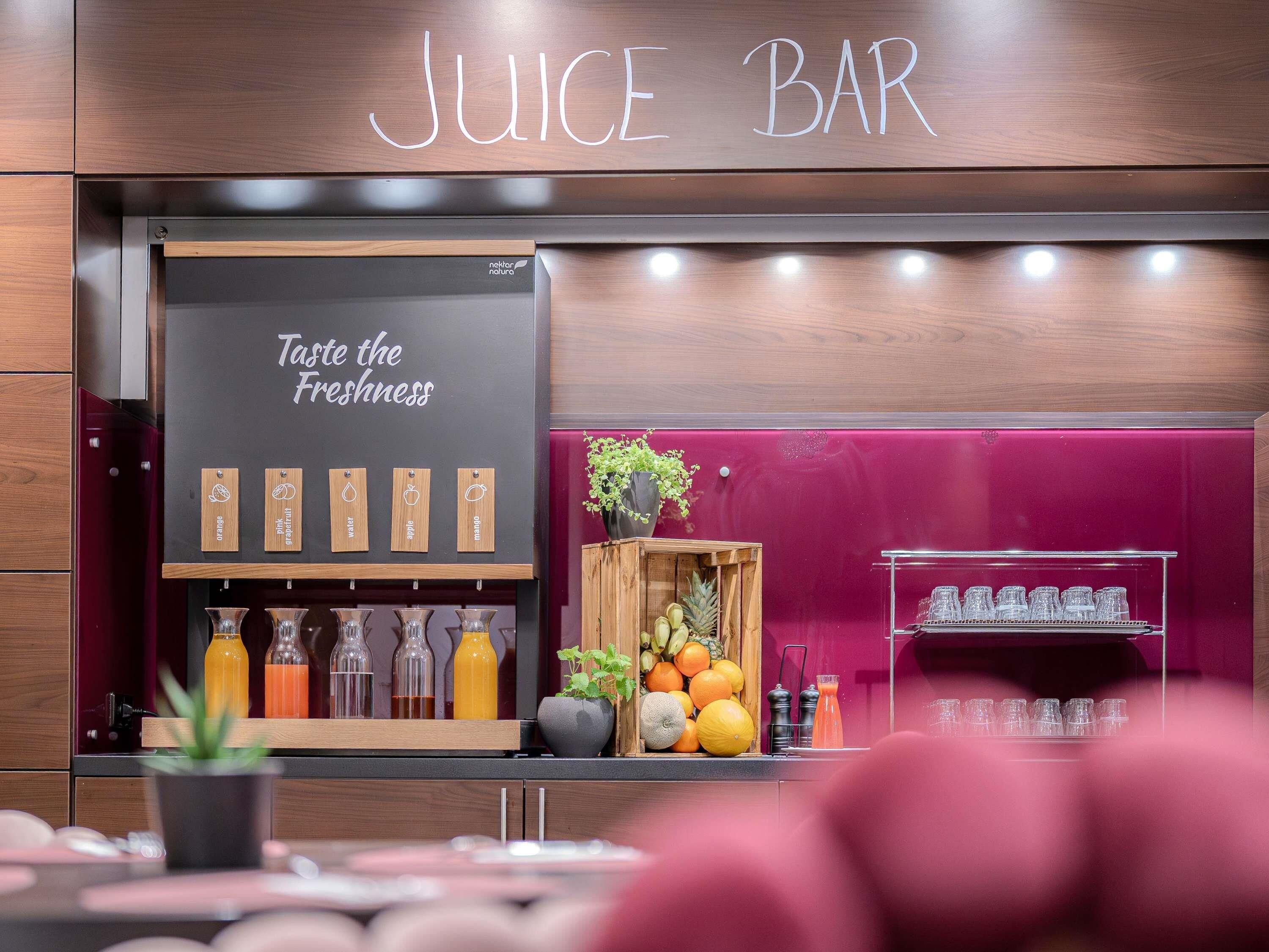 A juice bar with a sign reading 'Juice Bar' and 'Taste the Freshness.' There are various juice bottles and a wooden crate with fruits.