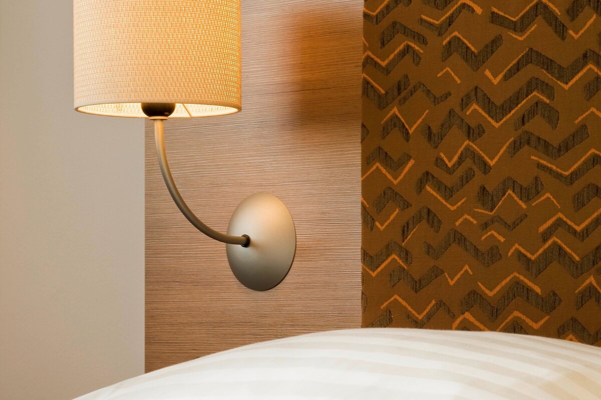 A modern lamp with a curved arm and a textured shade, positioned on a wall with a wooden panel and a patterned wallpaper.