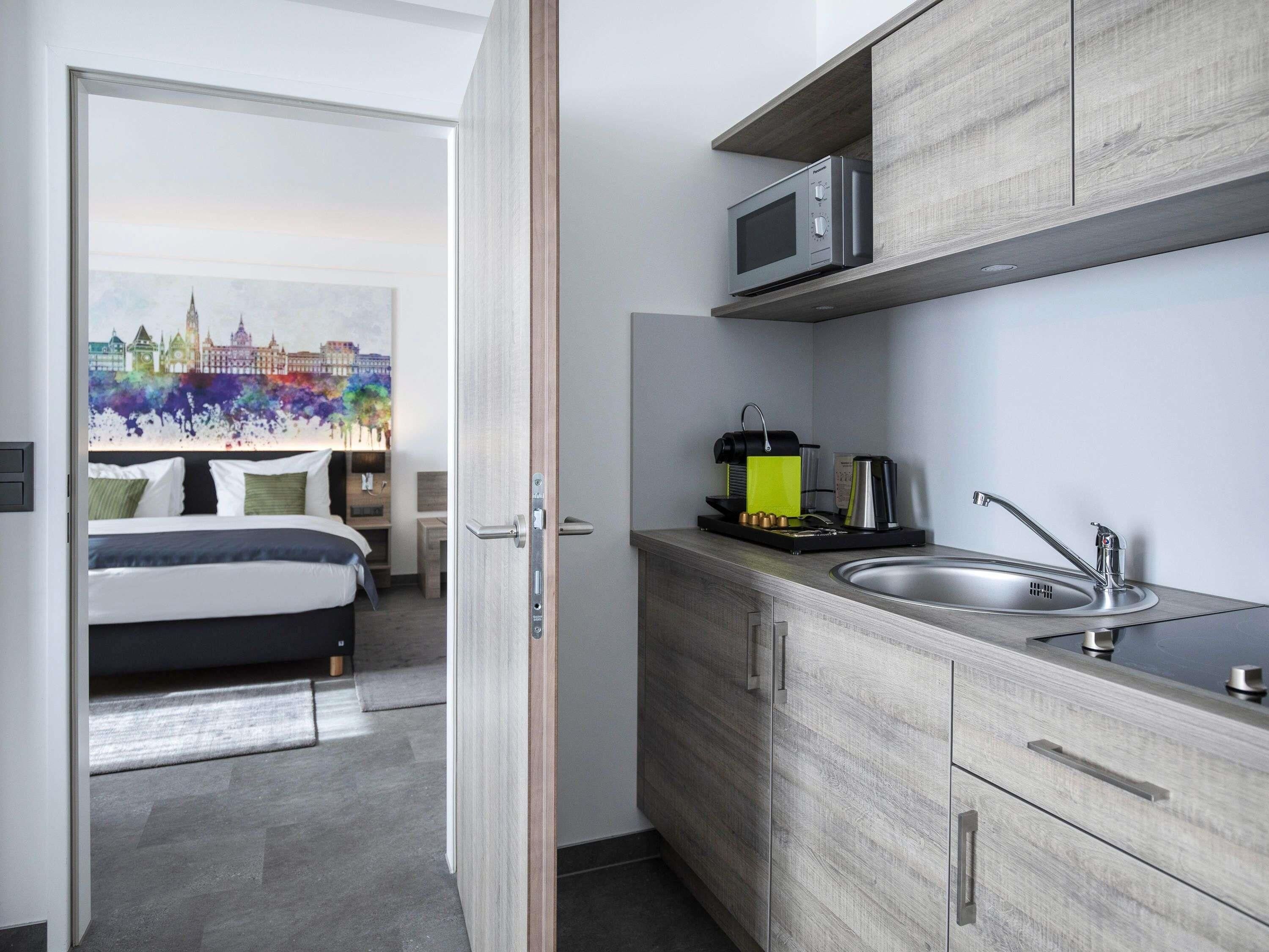 A compact hotel room with a bed, a kitchenette, and a cityscape painting on the wall.