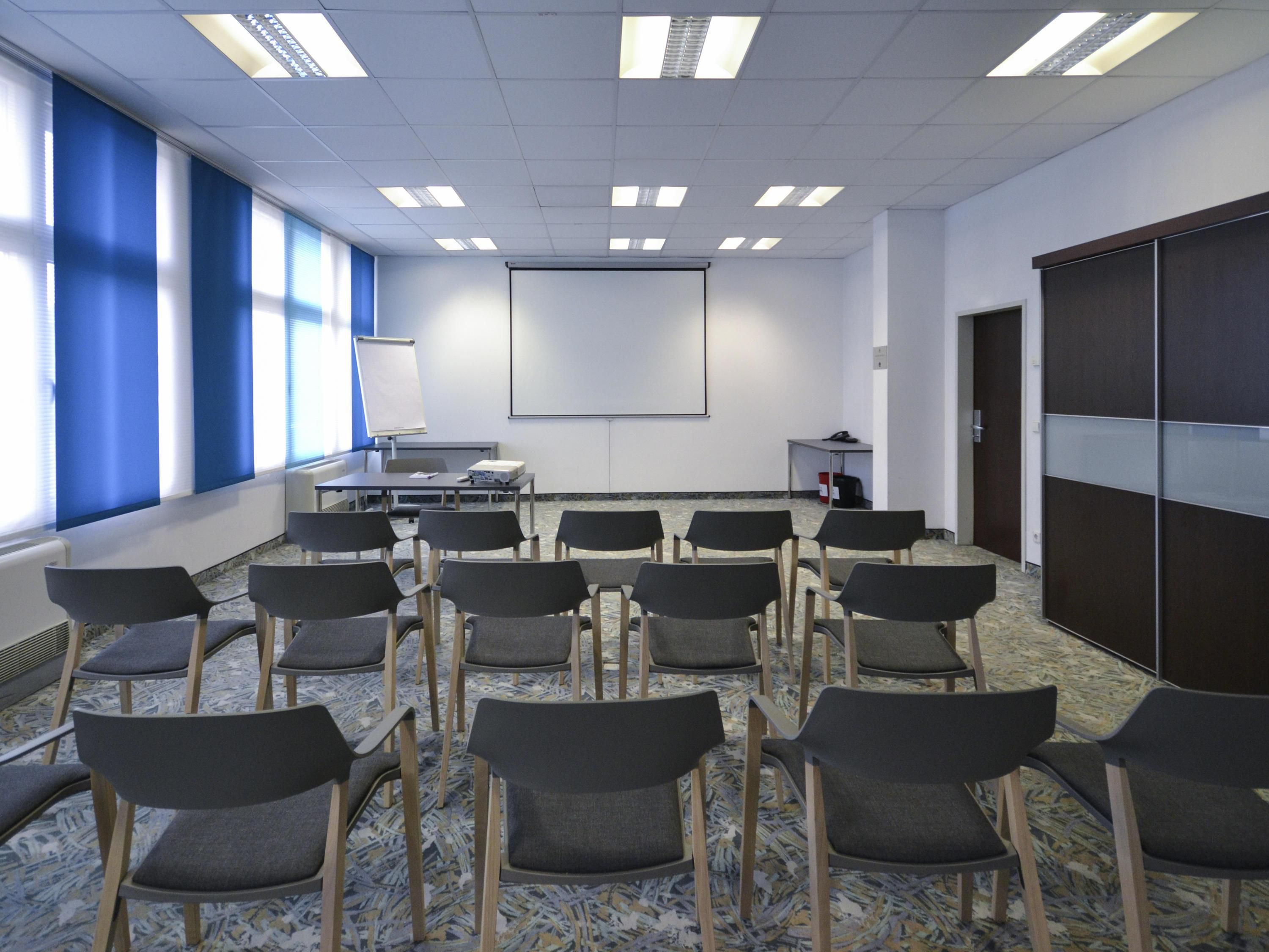 A conference room with rows of chairs, a projector screen, a whiteboard, and a table with a laptop.