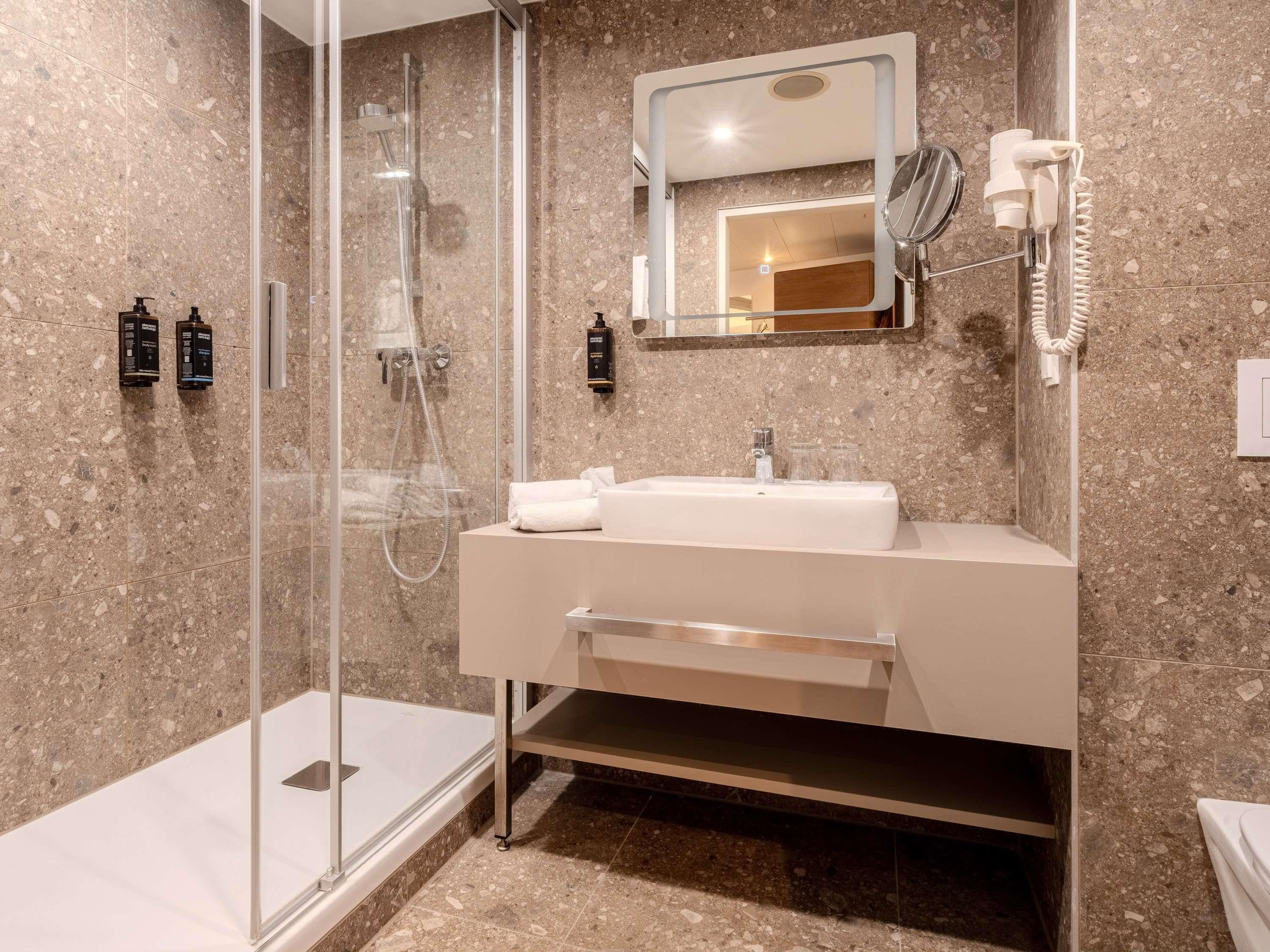 A modern bathroom with a glass-enclosed shower, a white sink with a mirror above it, and a hairdryer mounted on the wall.
