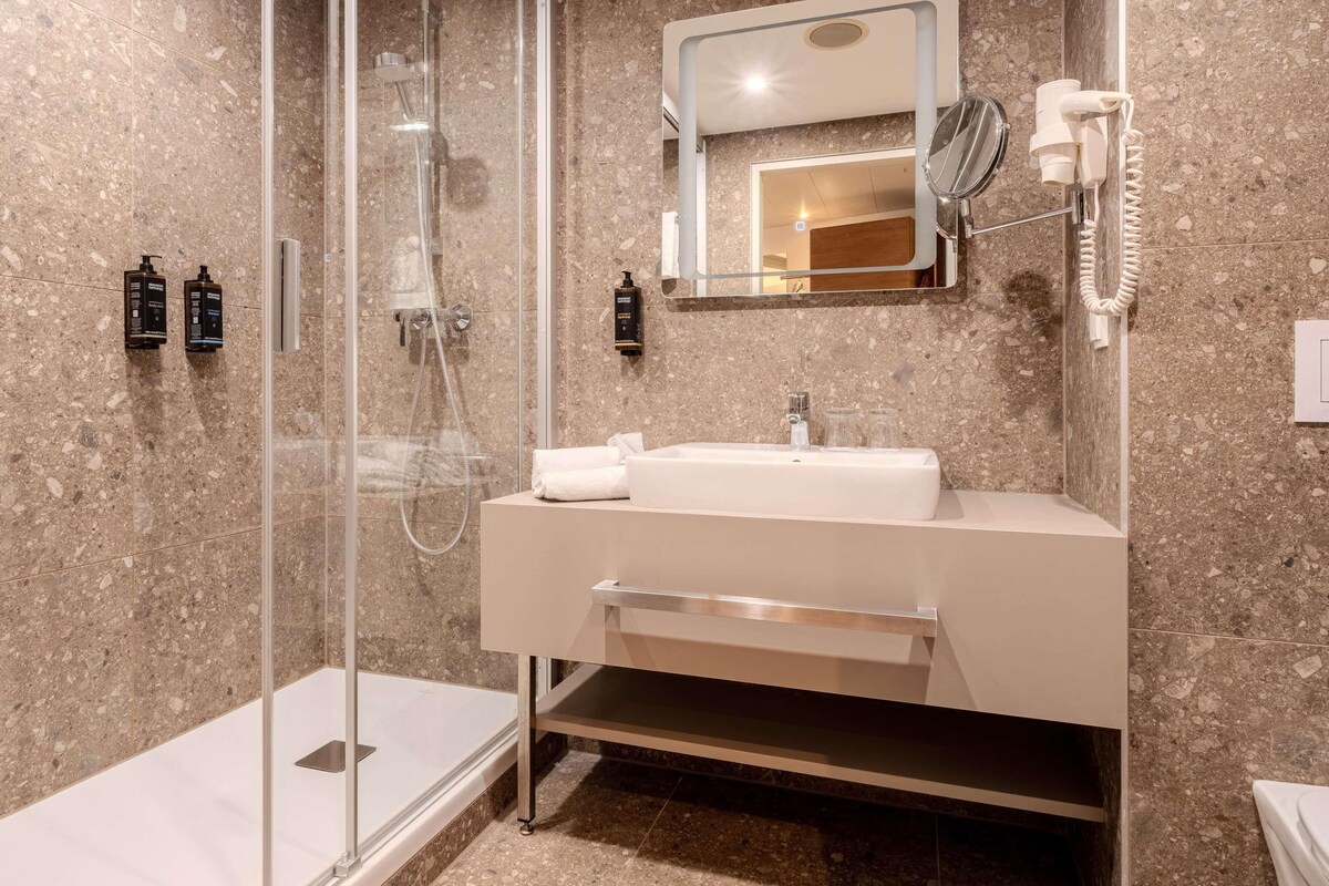 A modern bathroom with a glass-enclosed shower, a white sink with a mirror above it, and a hairdryer mounted on the wall.