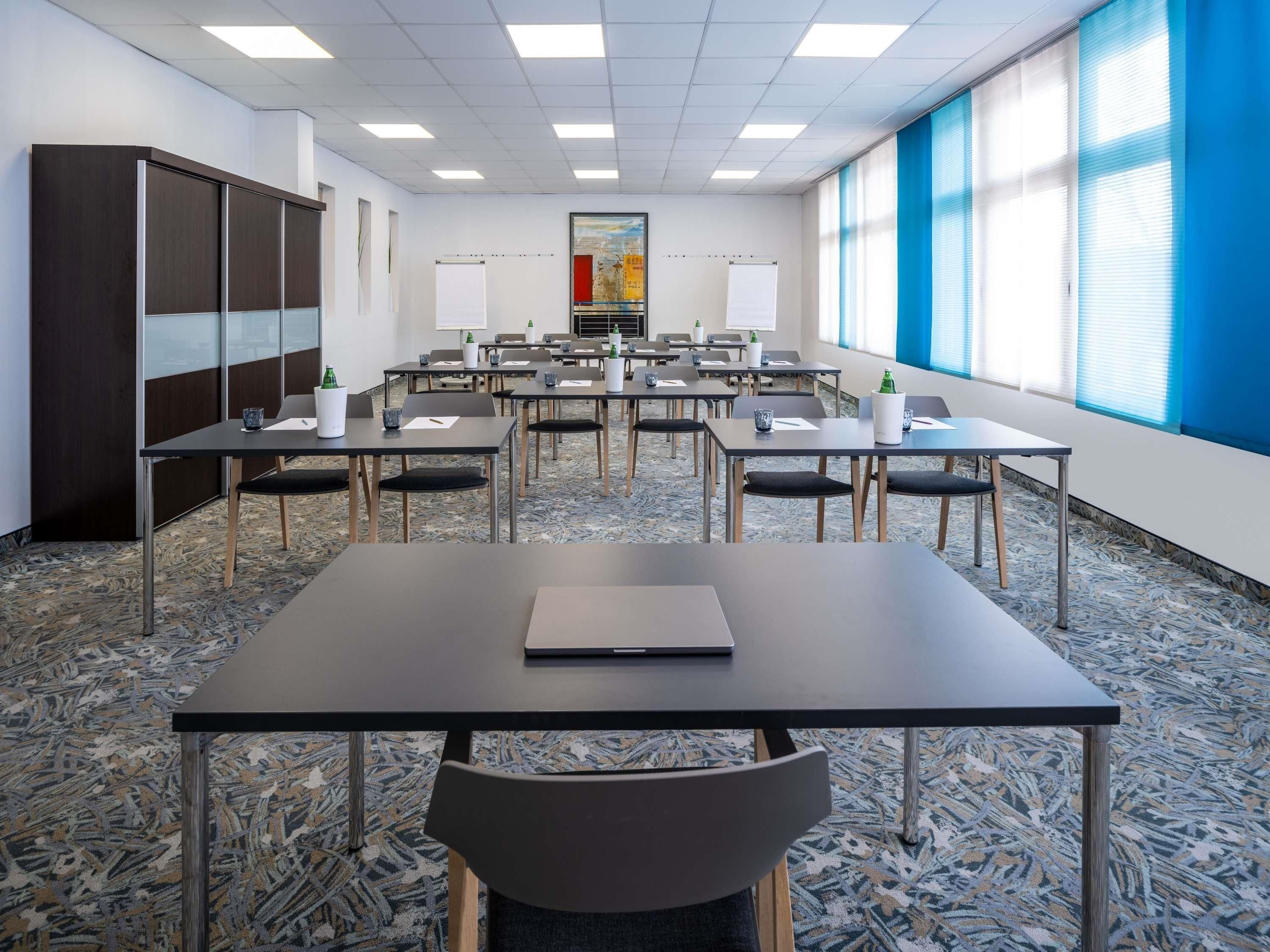 Une salle de conférence équipée de tables, de chaises et d’une grande fenêtre avec des stores.