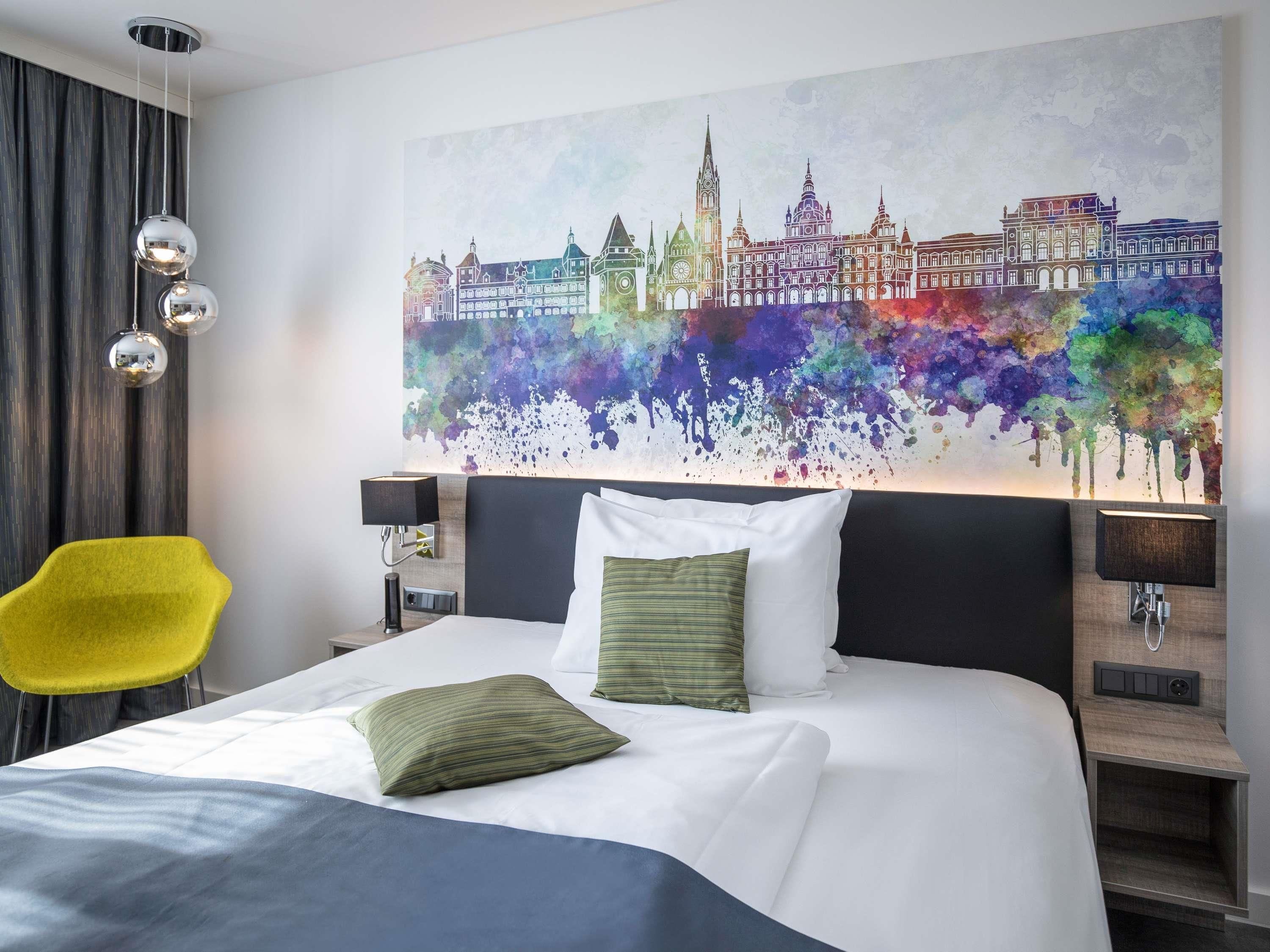 Quarto de hotel com uma cama, uma cadeira amarela, uma pintura grande com vista para a cidade e duas luminárias de cabeceira.