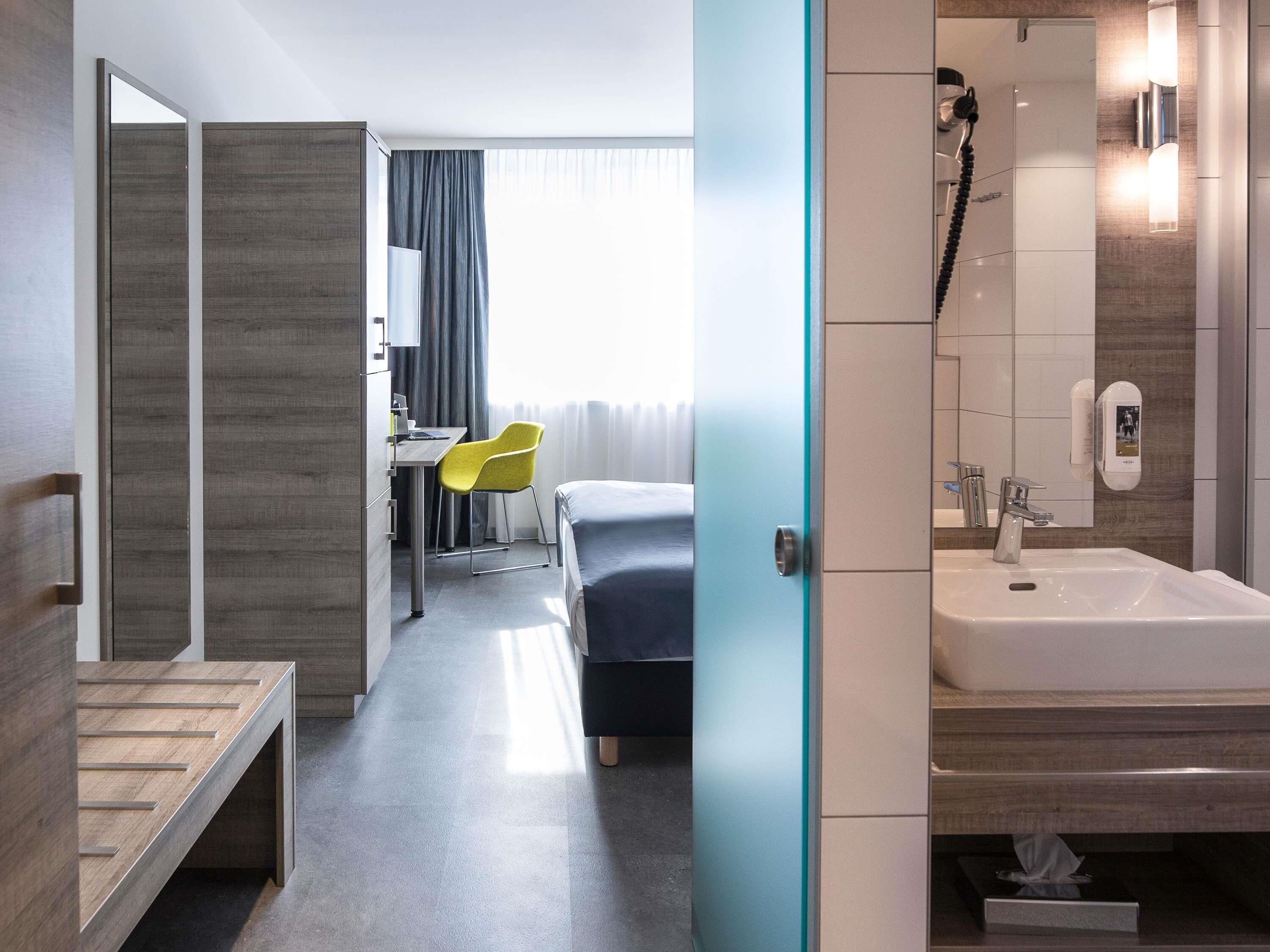 A hotel room with a bed, a desk with a chair, a bathroom with a sink and mirror, and a window with curtains.