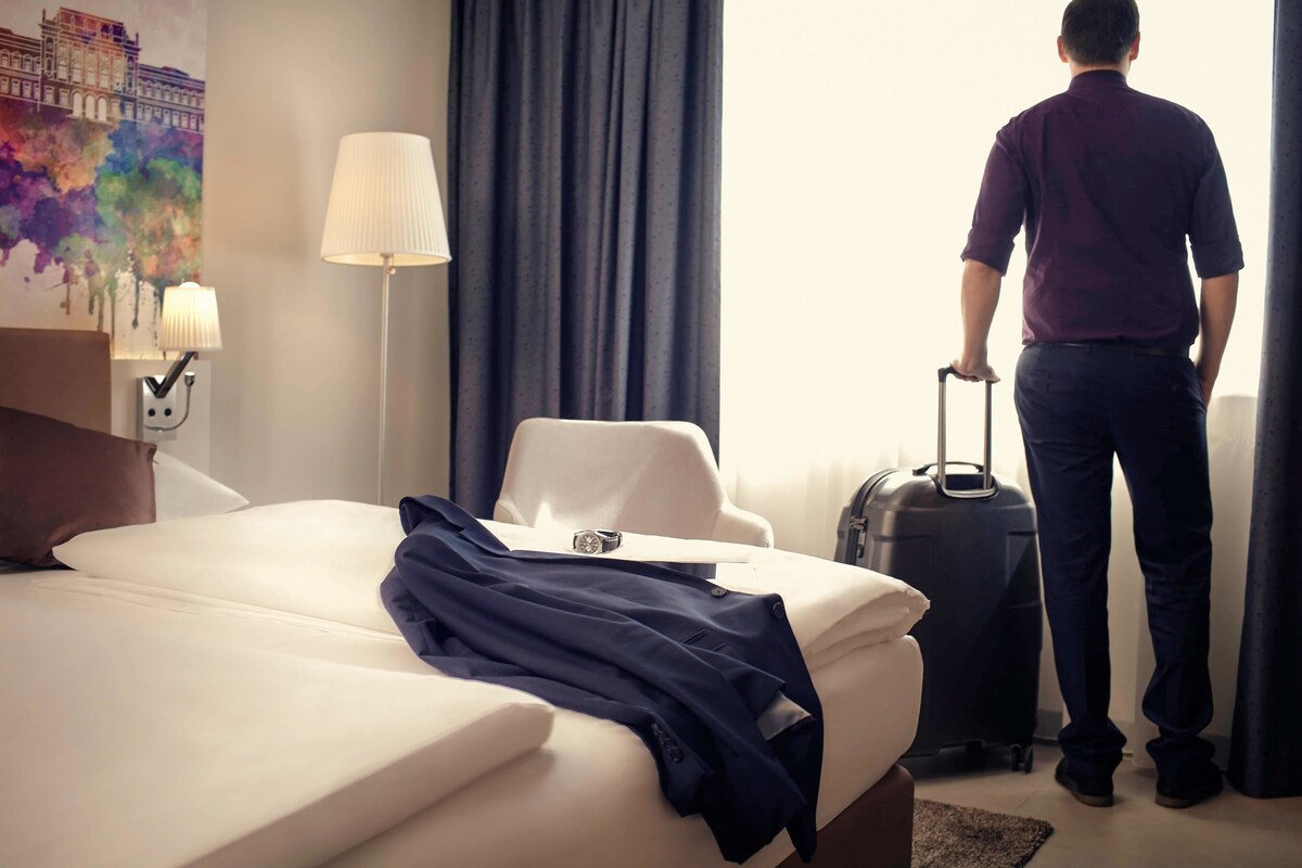 A hotel room with a bed, a lamp, a suitcase, and a painting on the wall.