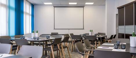 Un salón de conferencias con varias mesas, sillas, una pantalla de proyector y un amplio ventanal con persianas.