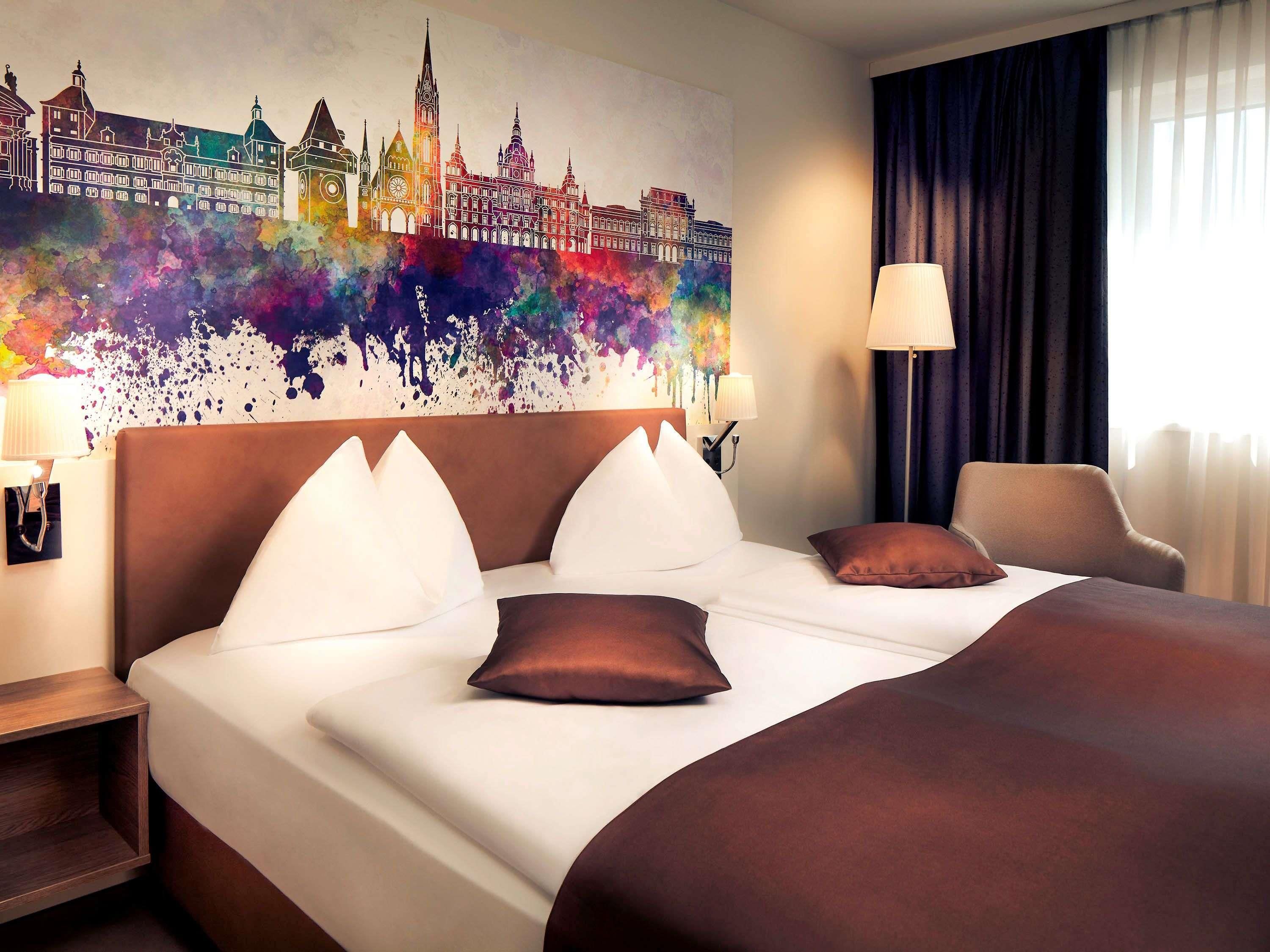 A hotel room with a large bed, a headboard, a nightstand, a lamp, a chair, and a colorful cityscape painting on the wall.