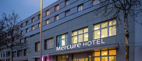 Un edificio moderno de hotel con un llamativo letrero de 'Mercure HOTEL', ventanas amplias y una entrada bien iluminada.