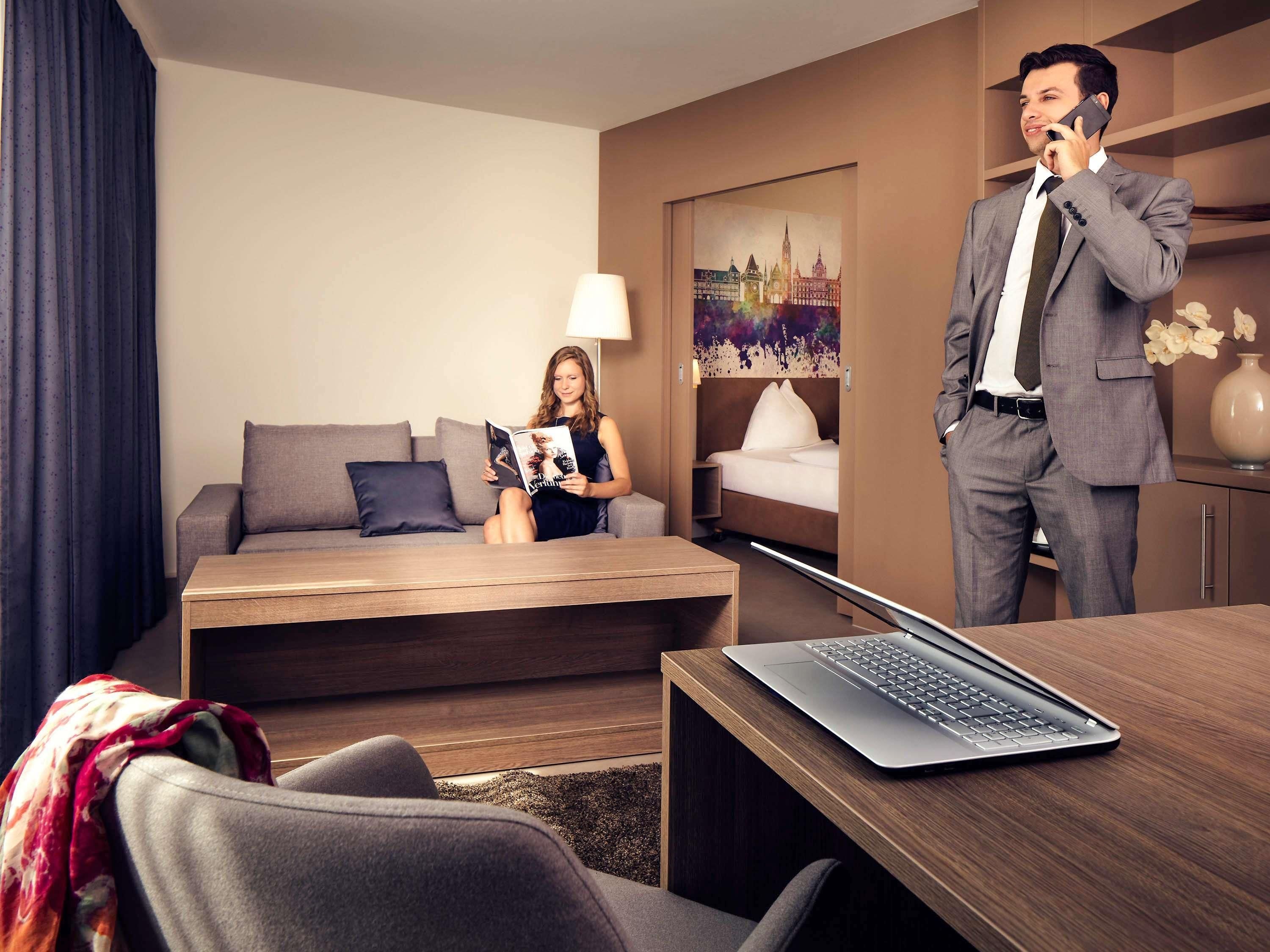 Une chambre d’hôtel moderne, équipée d’un canapé, d’un lit, d’un bureau avec un ordinateur portable, et où une personne est en train de téléphoner.