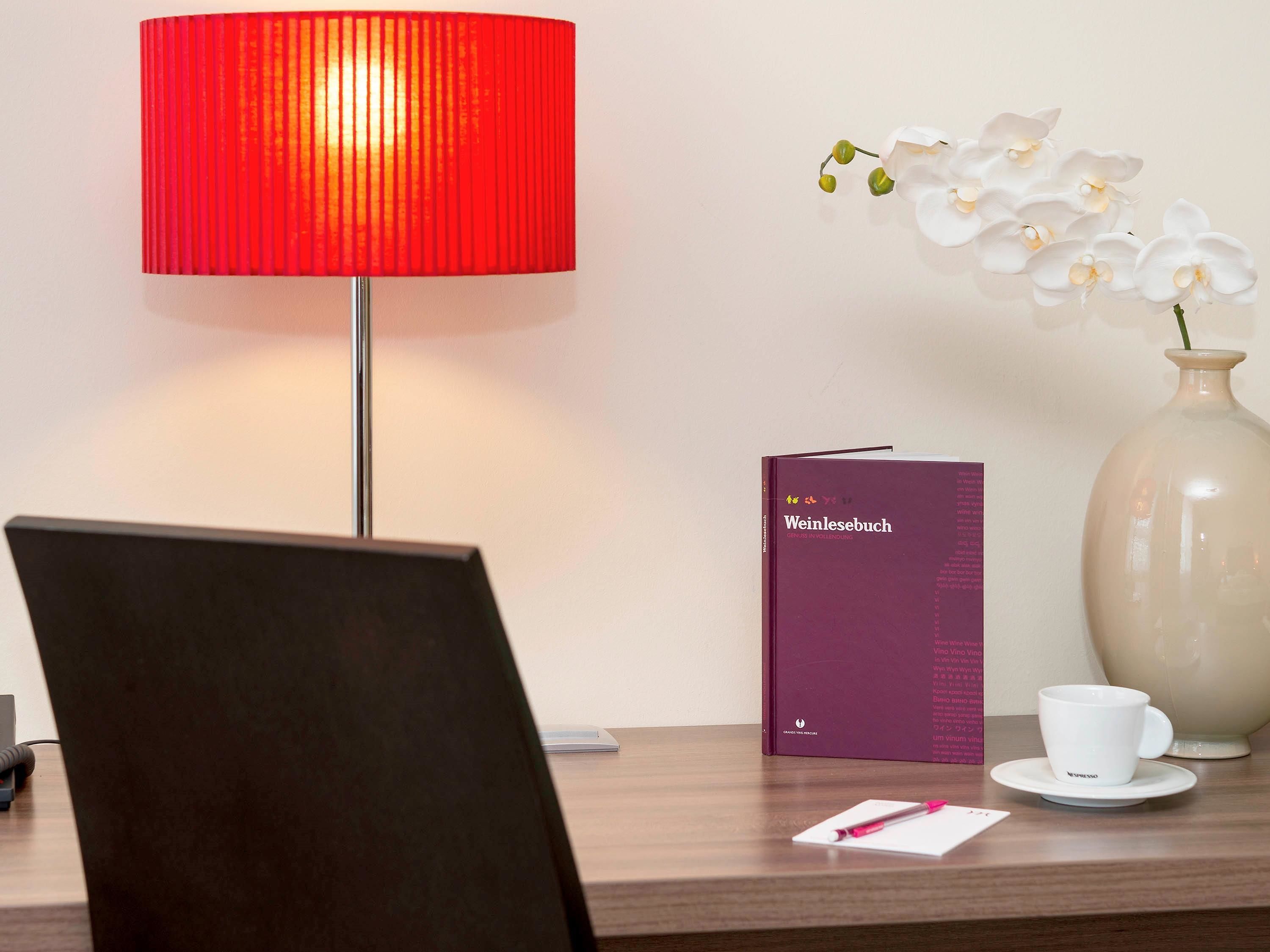 Un bureau avec une lampe rouge, un livre intitulé « Weinlesebuch », une tasse blanche et un vase contenant des orchidées blanches.